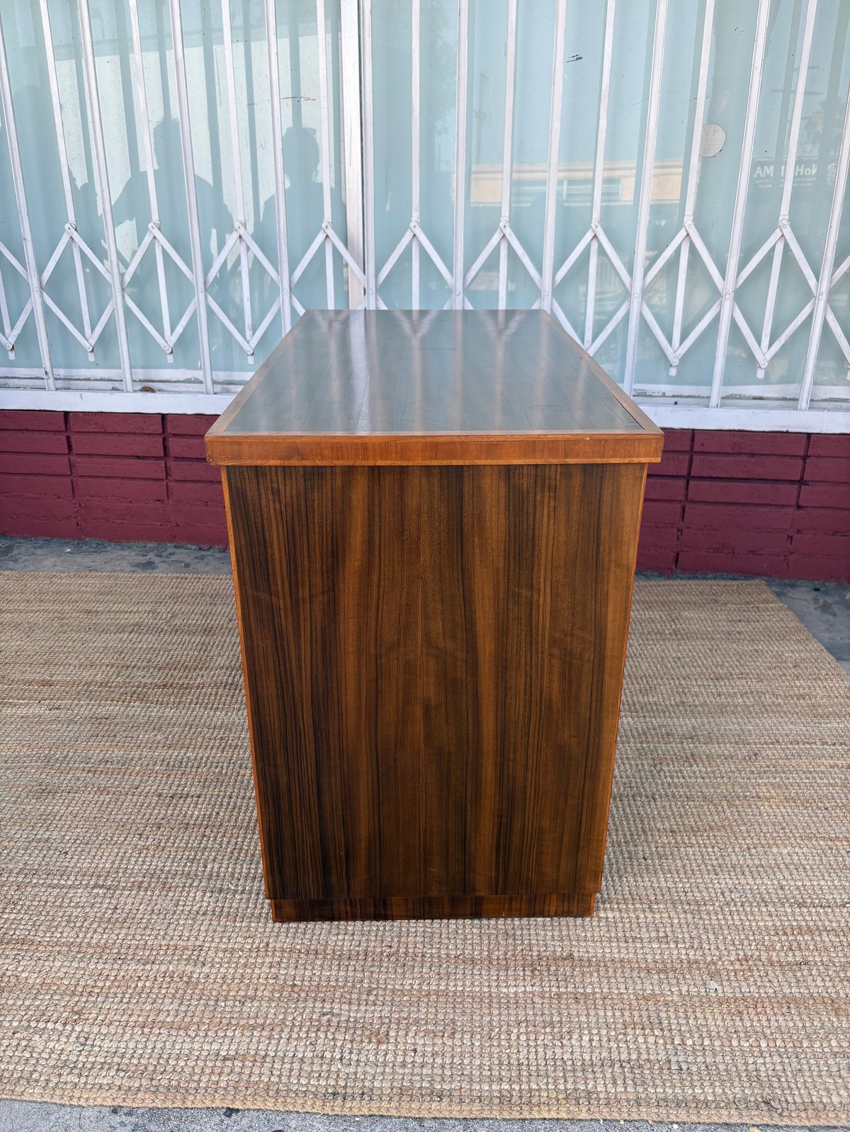Wooden pedestal table with dark top on paved outdoor surface against white accordion gate and burgundy wall