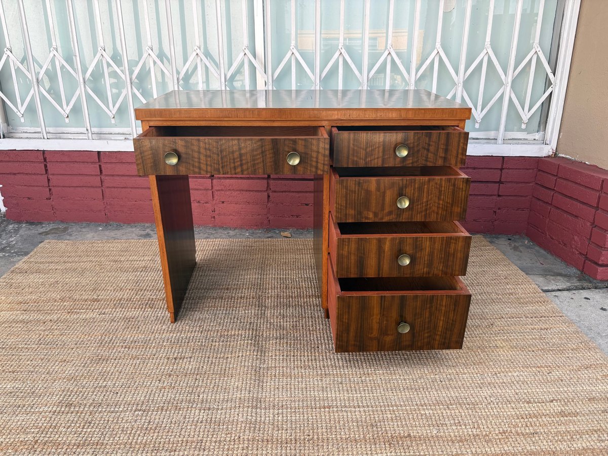 Wooden desk with five drawers and a side panel, positioned on a woven mat against a red brick wall and white window.