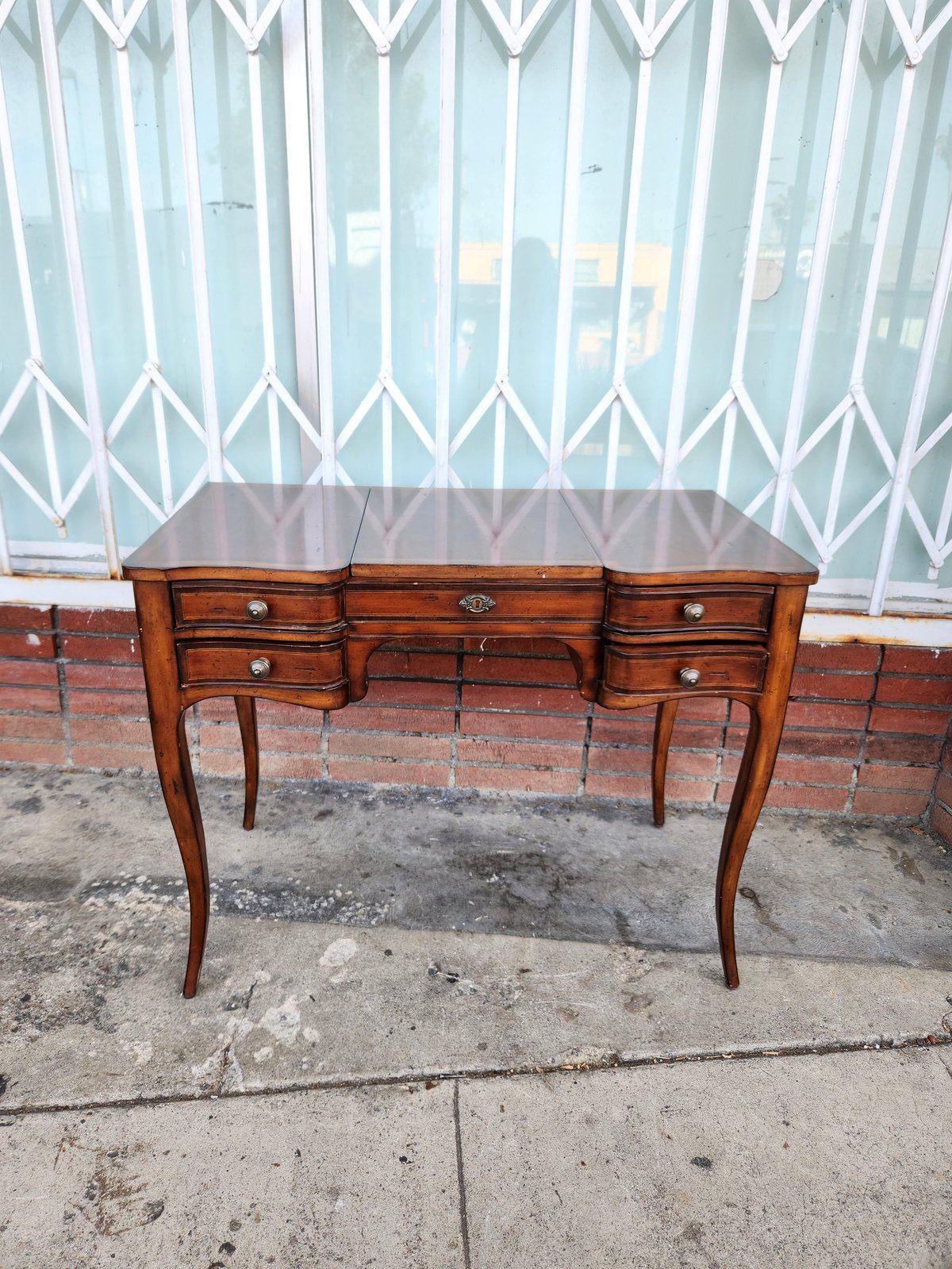 Vintage wooden vanity desk with drawers and curved legs against a white metal security gate