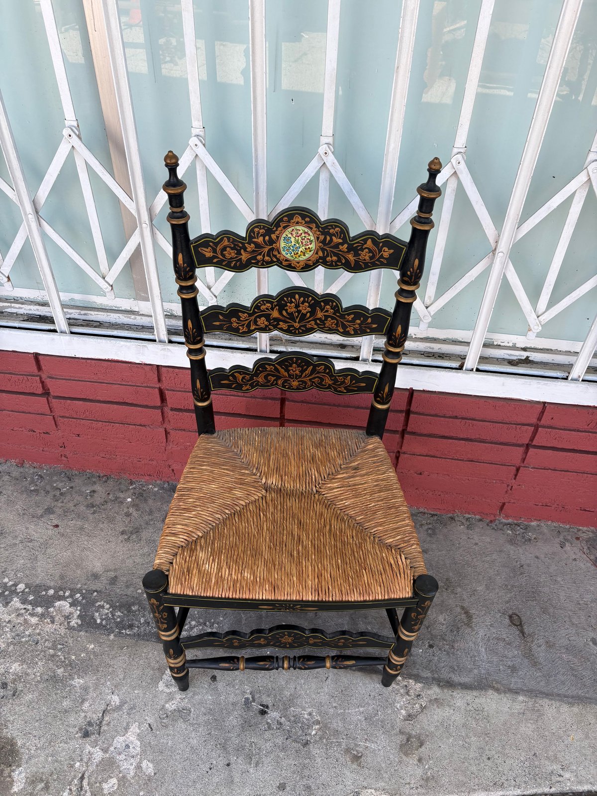 Ornate black wooden chair with woven rush seat and decorative gold details, photographed outdoors against a red brick wall with white latticed window backdrop