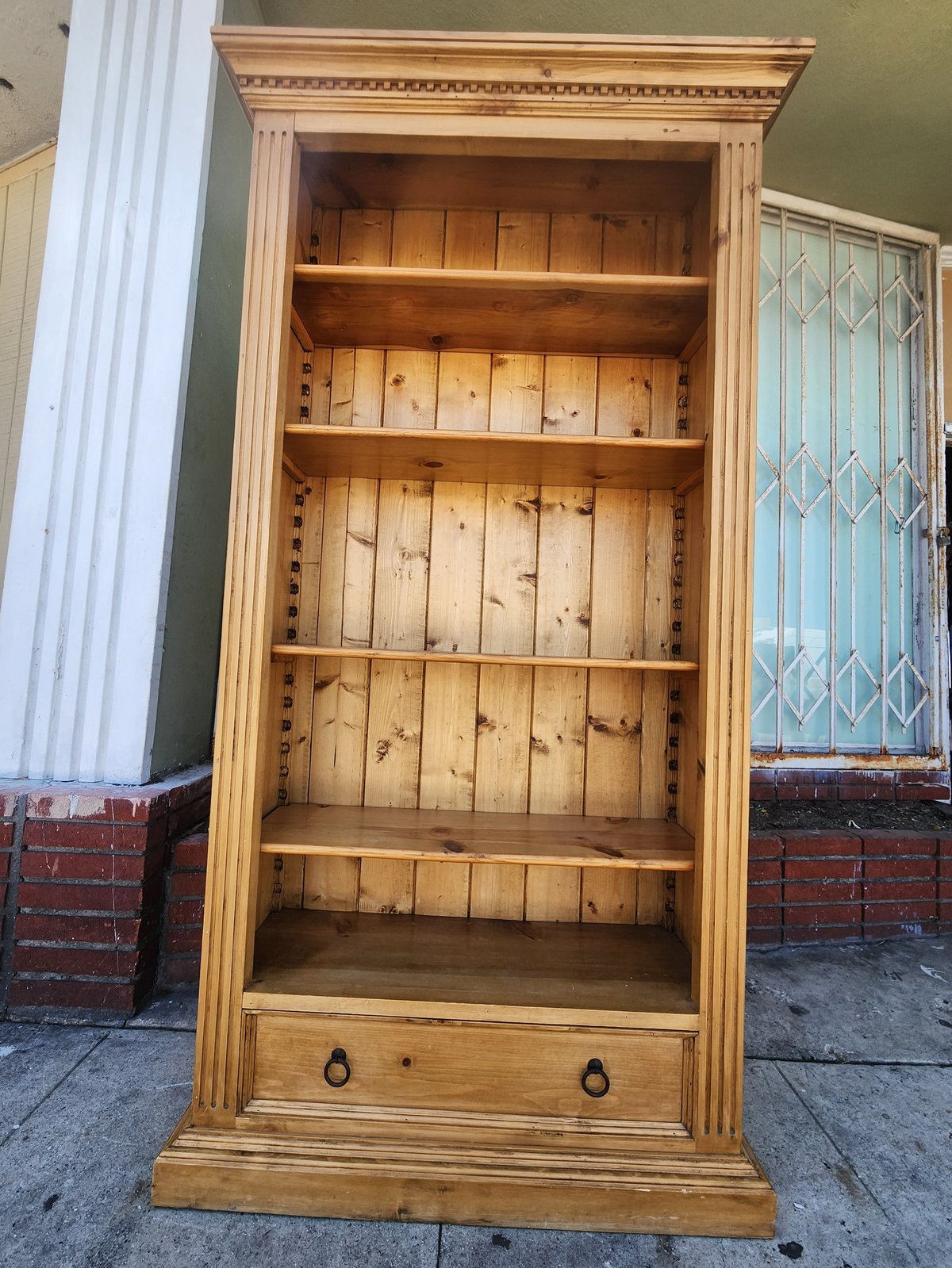 Tall wooden bookcase with five shelves and bottom drawer, displayed outdoors against a green wall and brick base