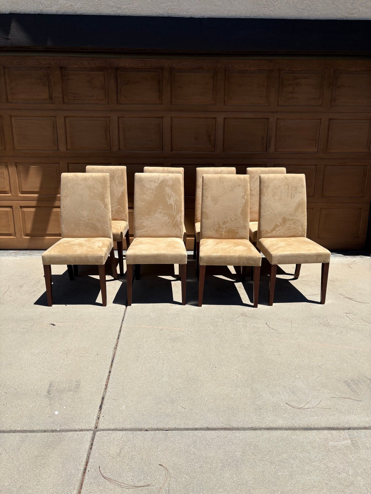 Set of four upholstered dining chairs with beige fabric and wooden legs arranged on a concrete driveway in front of a wooden garage door.