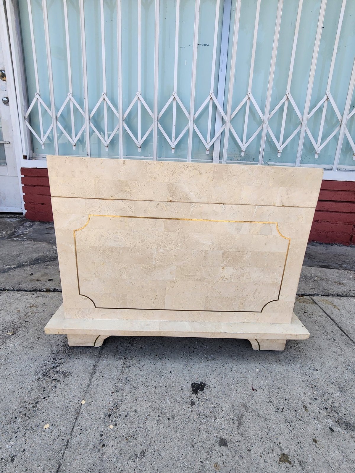 Cream-colored wooden cabinet with decorative panel sitting on concrete in front of corrugated metal gate