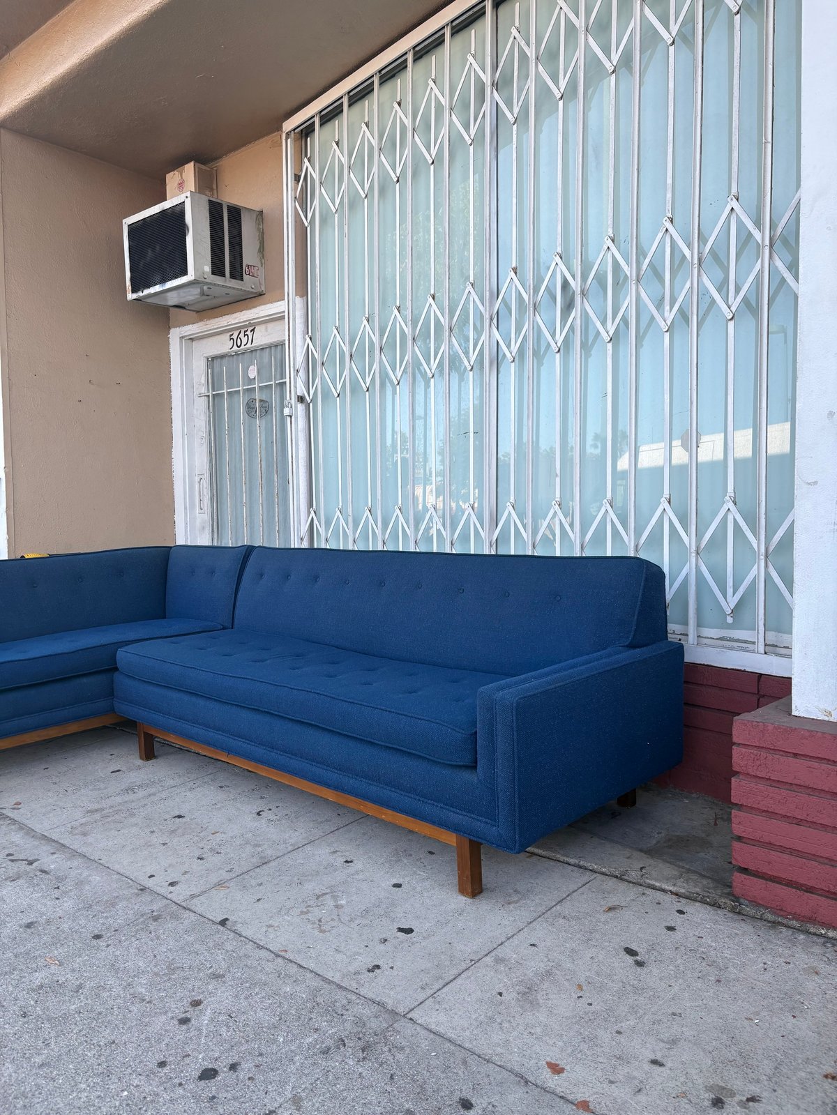Blue mid-century modern sectional sofa on a concrete porch with a white accordion gate, air unit, and red steps