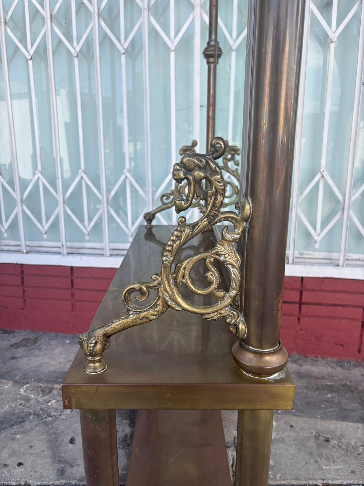 Ornate brass dragon sculpture mounted on a pole with decorative scrollwork detail, displayed outdoors against a white metal gate and red wall