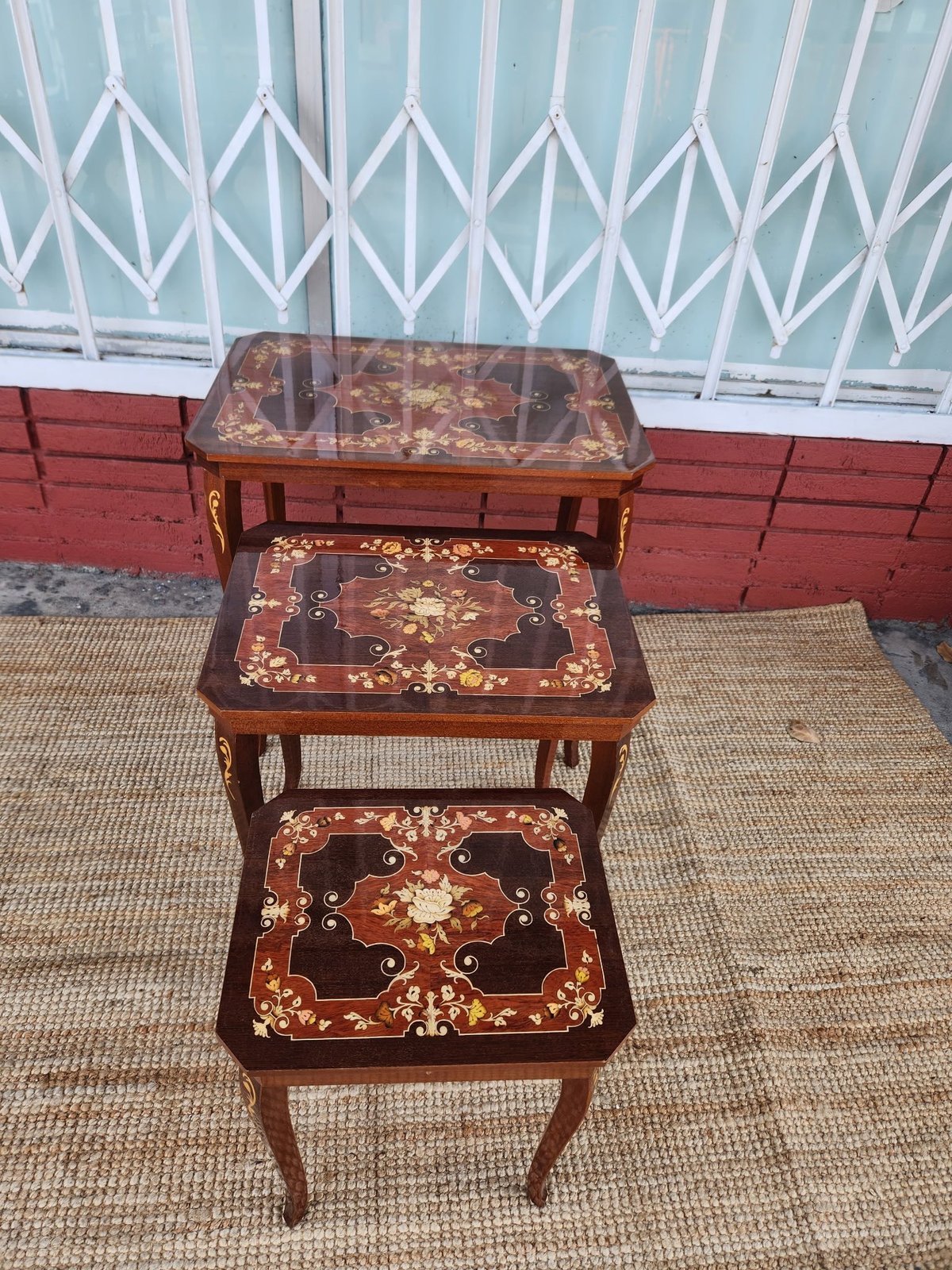 Three wooden nesting tables with ornate inlaid floral designs displayed outdoors against a red and white garage door.