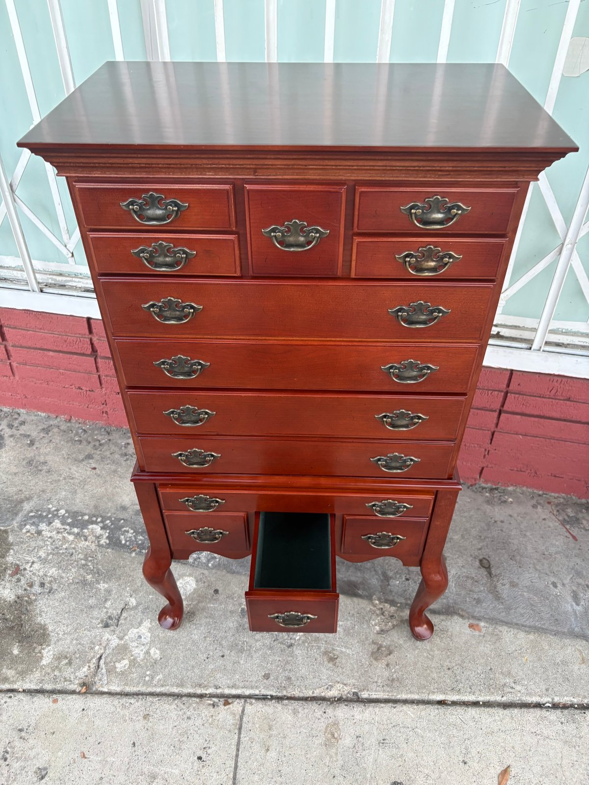 Tall red wood dresser with brass handles and cabriole legs sits on pavement against light blue storefront.