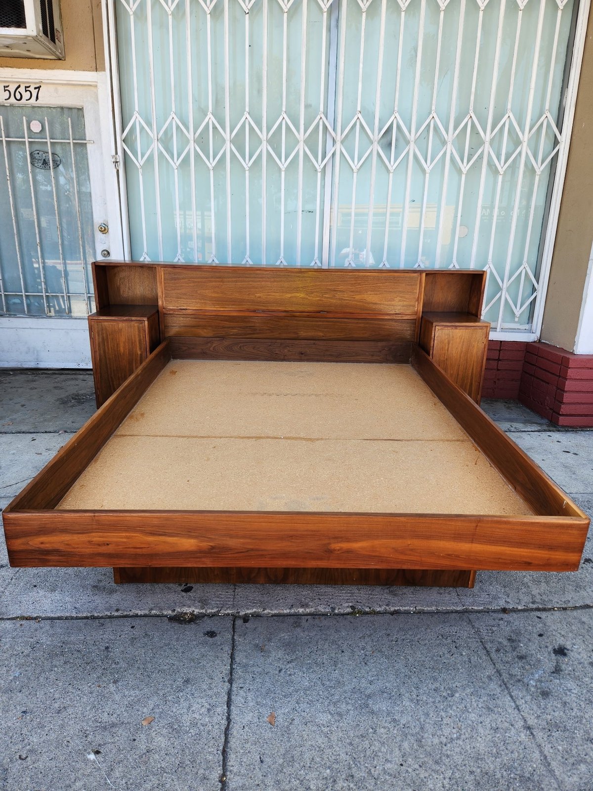 Wooden platform bed frame with beige mattress displayed on outdoor pavement