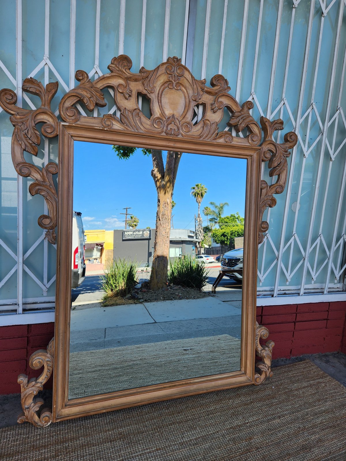 Ornate wooden framed mirror with scrolled details leaning against a metal gate, reflecting palm trees and buildings