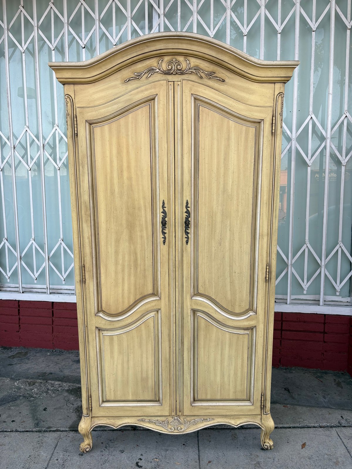 Cream-colored ornate wooden armoire with curved top, decorative carvings, and cabriole legs against metal gate backdrop