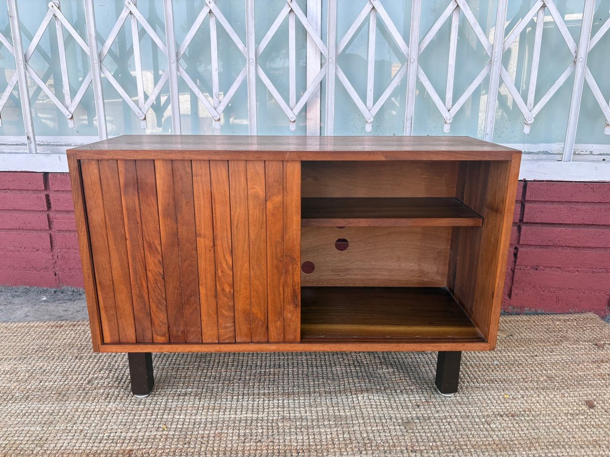 Mid-century modern wooden cabinet with sliding door open, showing two shelves inside, against a brick and lattice fence background