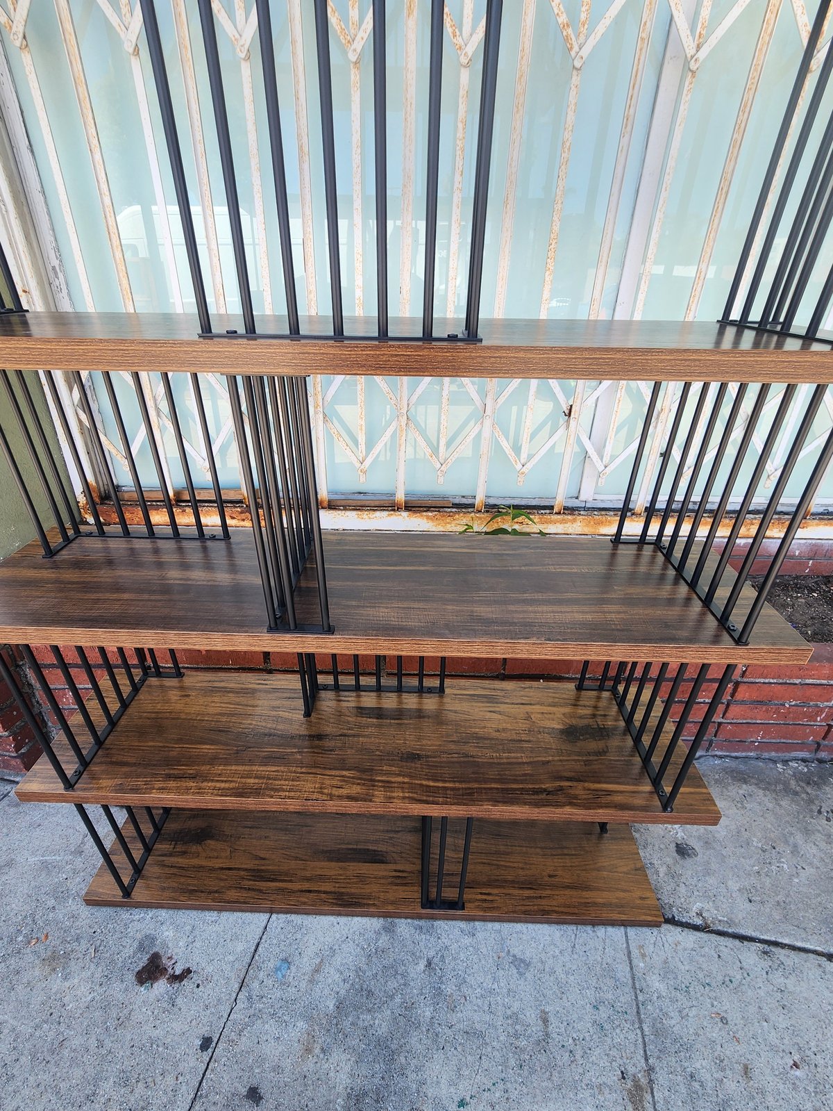 Wooden shelving unit with four horizontal shelves and black metal spindle dividers, displayed outdoors against a building