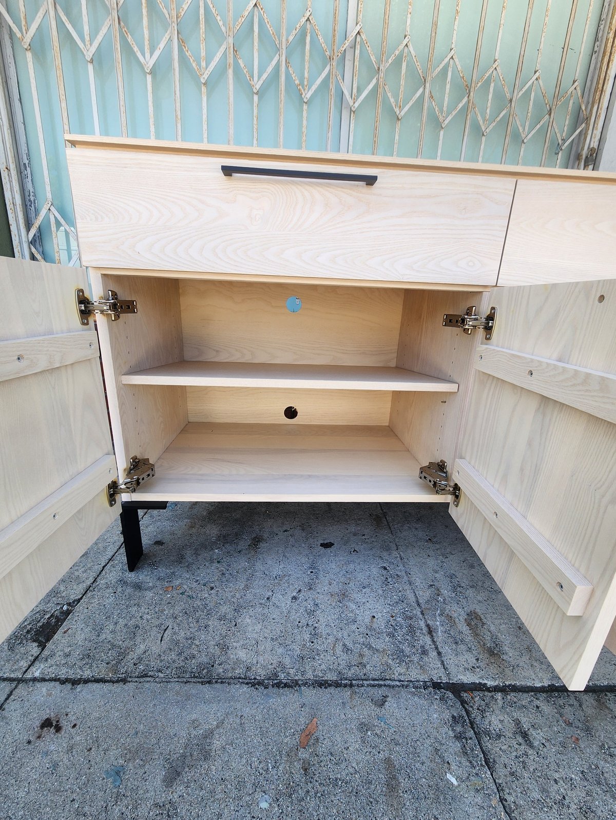 Open light wood cabinet with two interior shelves and geometric patterned backing, mounted above concrete floor