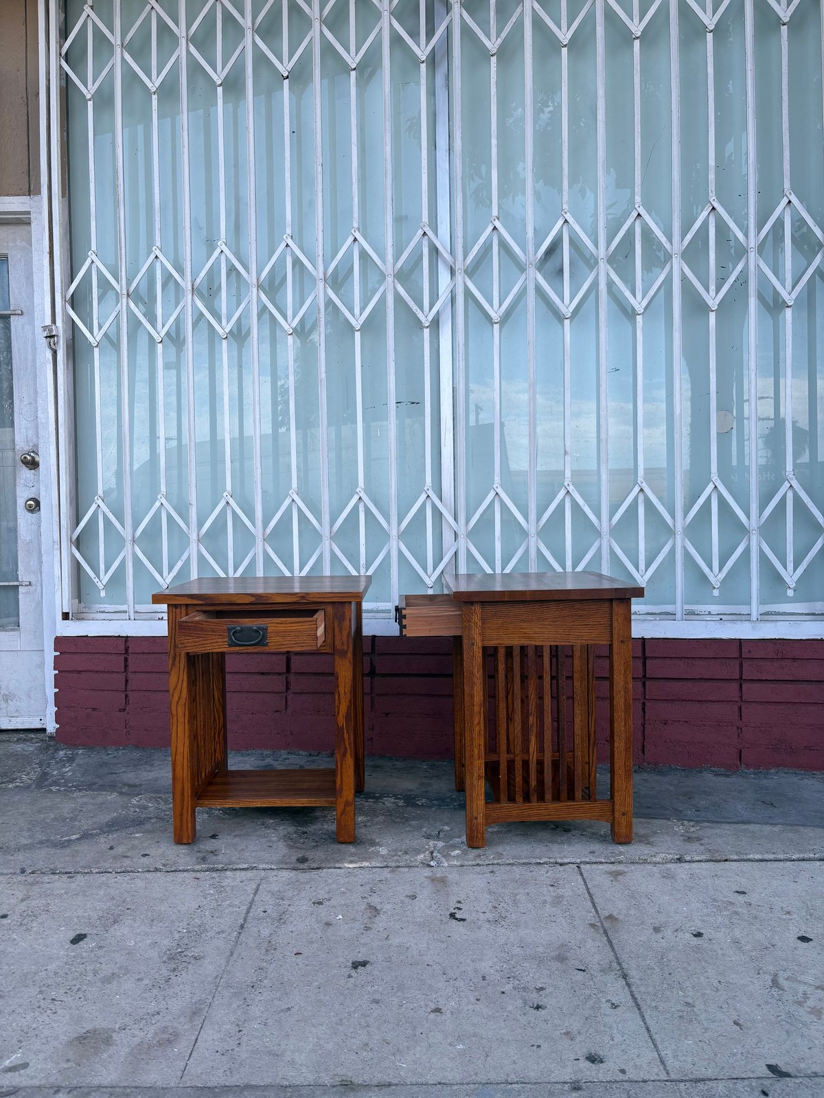 Two wooden side tables on a sidewalk in front of a white metal security gate and red brick wall