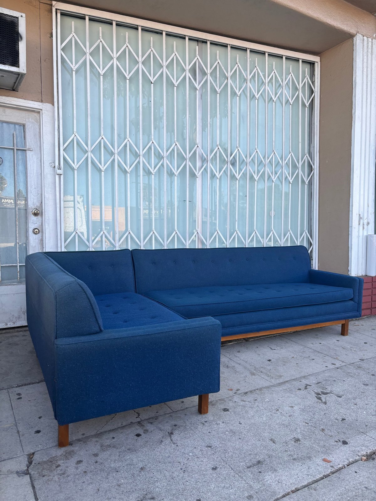 Blue mid-century modern sectional couch on a sidewalk in front of a storefront with metal security gate
