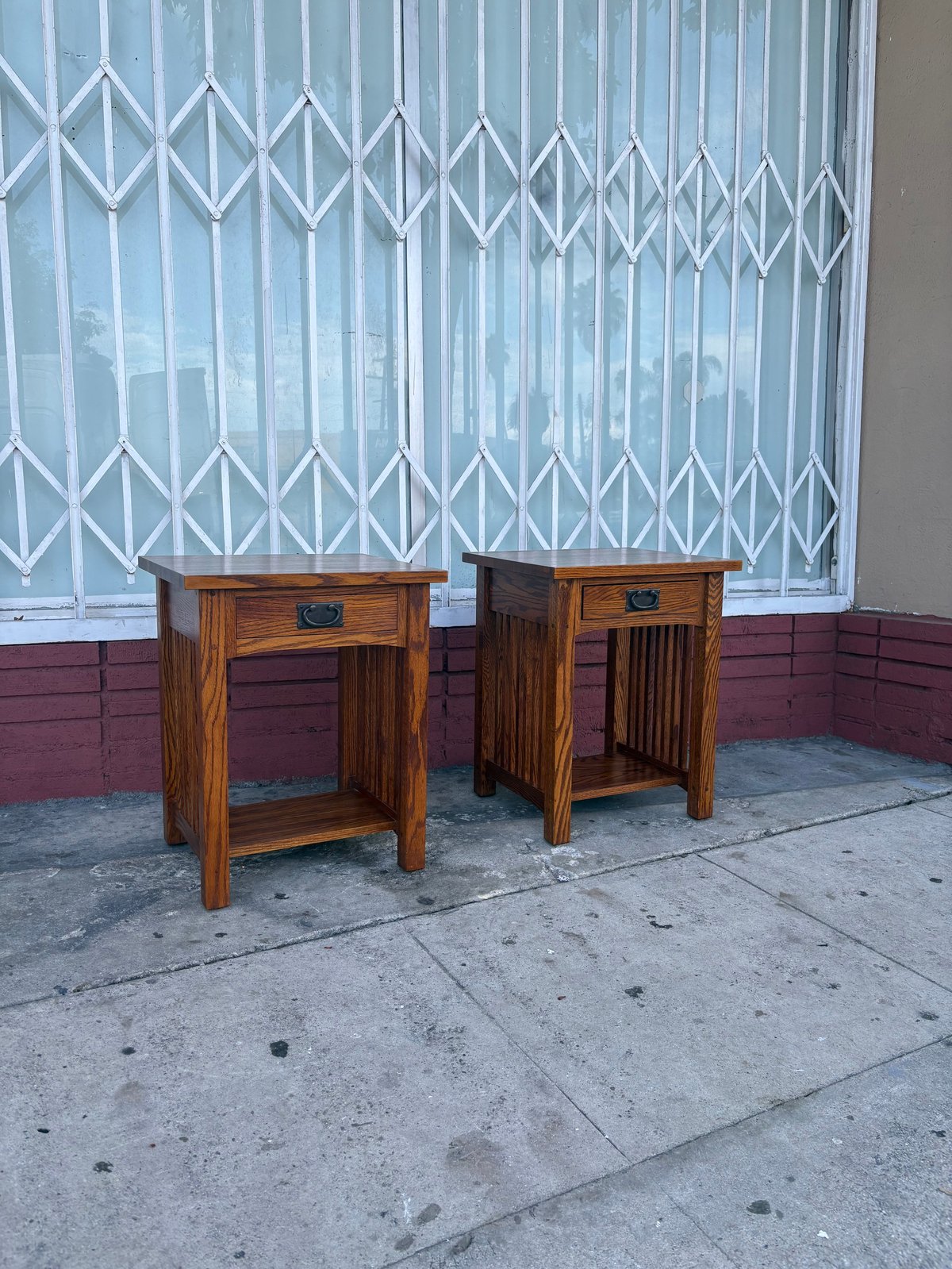 Two wooden nightstands with drawers displayed on a sidewalk in front of a metal accordion gate and red brick wall