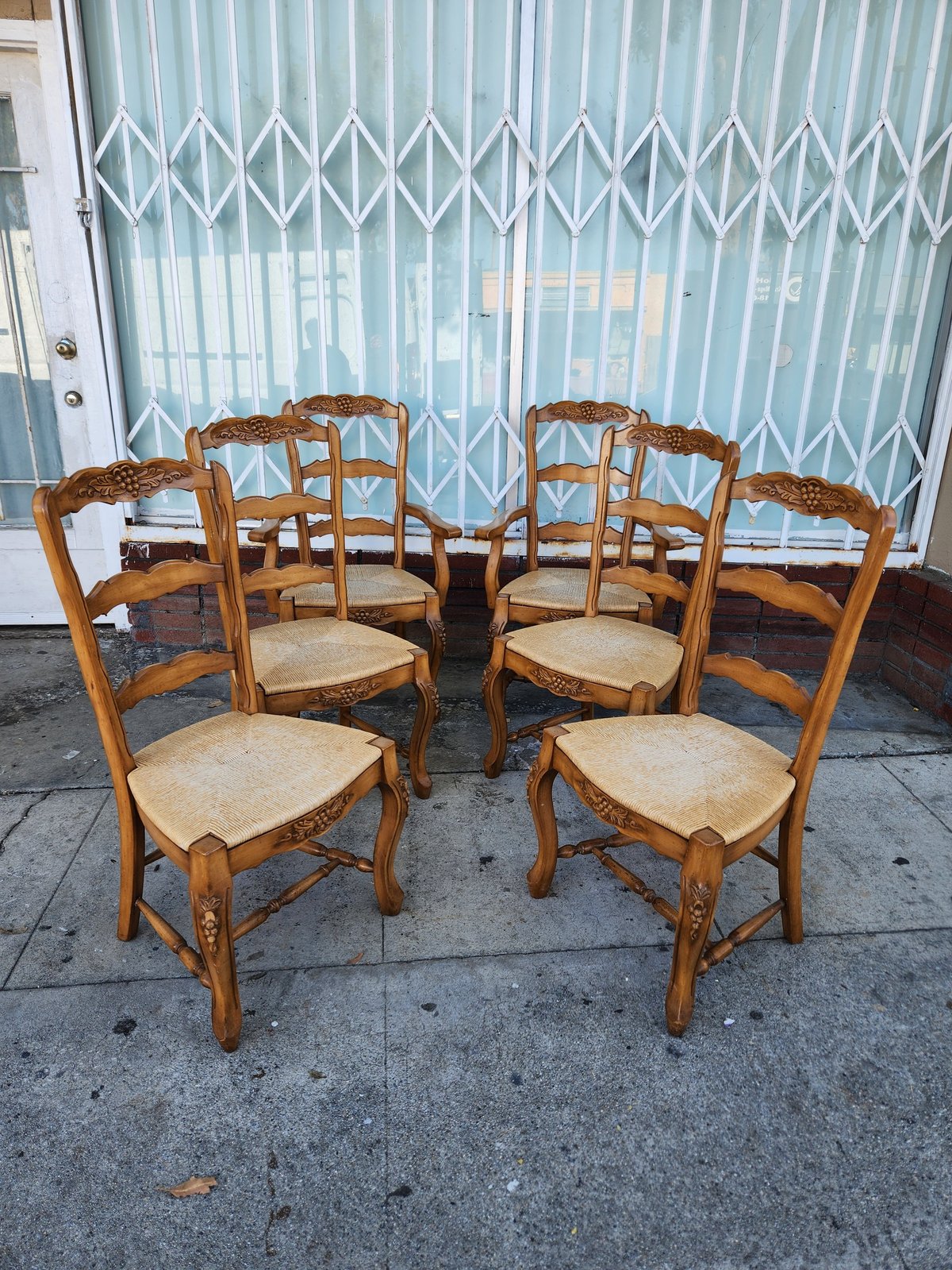 Four wooden chairs with woven rush seats arranged in a circle on a concrete surface in front of a light blue corrugated metal gate
