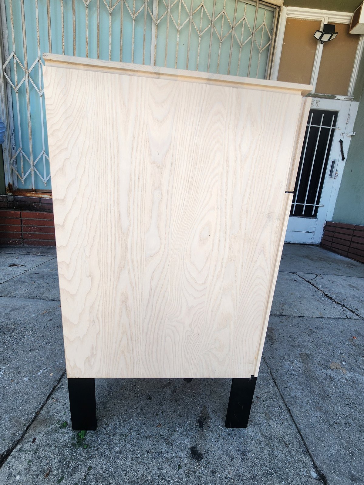 Plywood cabinet with black legs on concrete, displayed against residential building with blue-painted gate