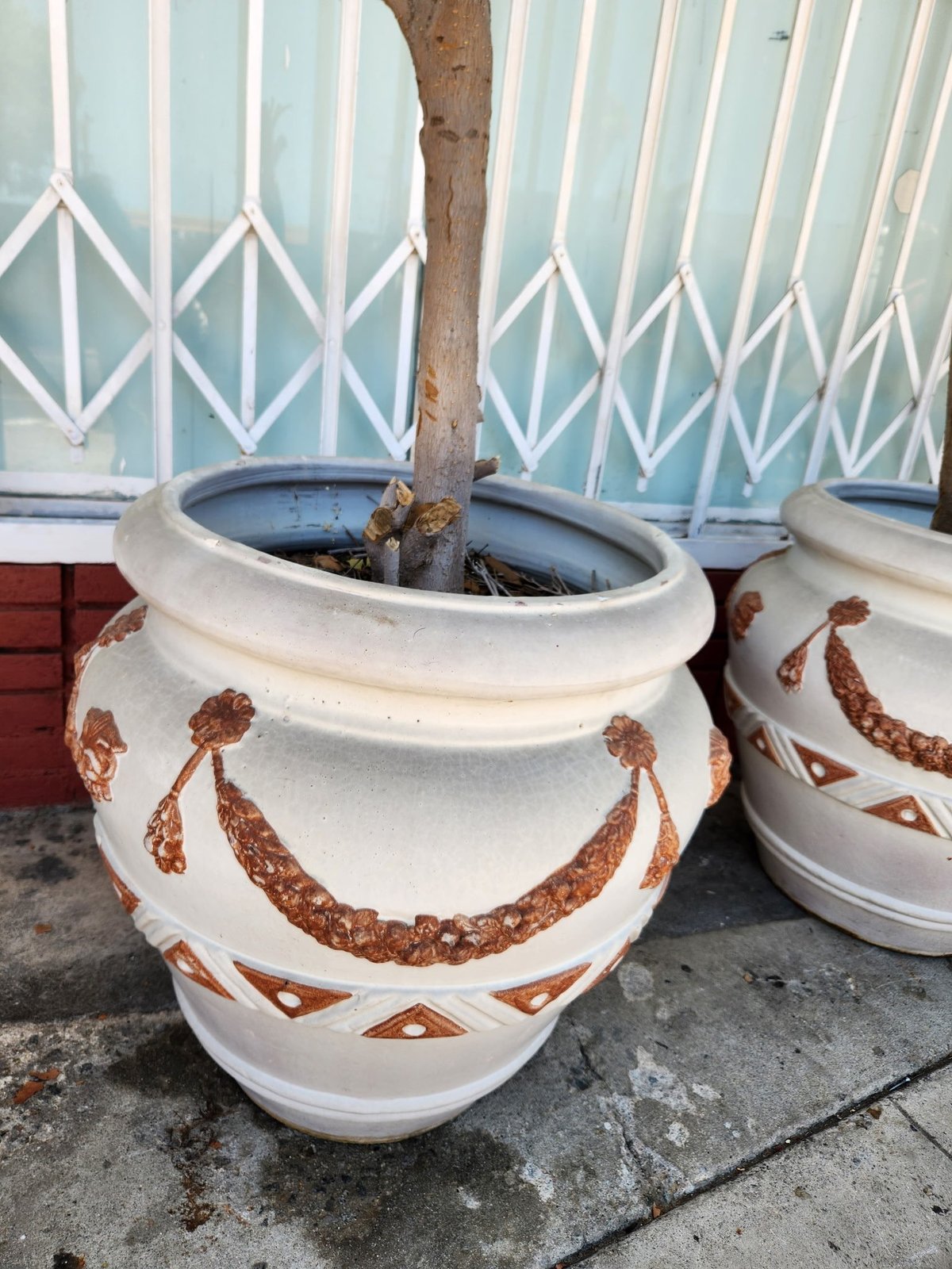 Large decorative white ceramic tree planter with russet-brown geometric patterns, holding a small tree trunk, positioned against a light blue striped metal fence