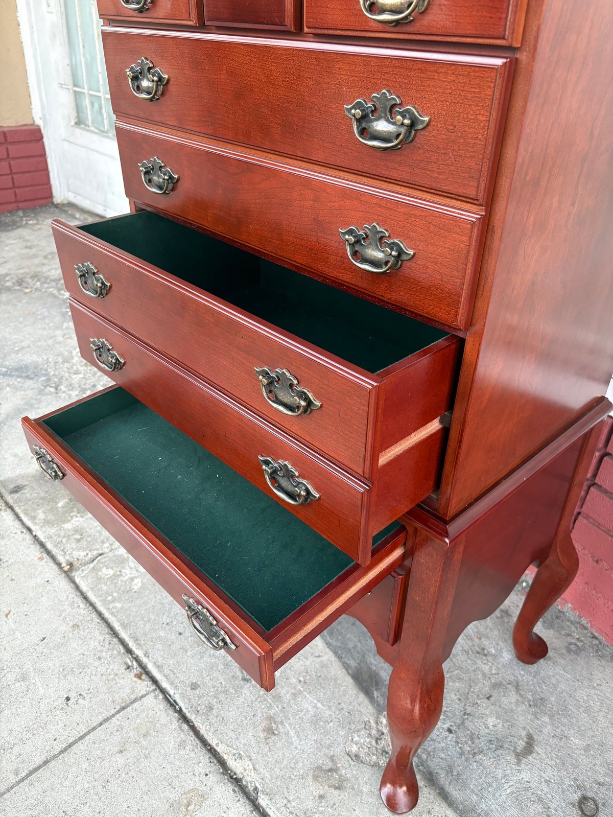 Red wooden dresser with ornate handles and two open drawers revealing green felt lining, displayed outdoors on concrete pavement