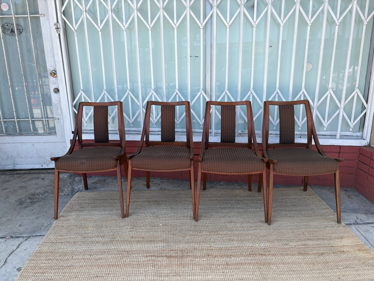 Four mid-century wooden dining chairs with vertical slat backs arranged in a row on a beige rug against a white metal gate
