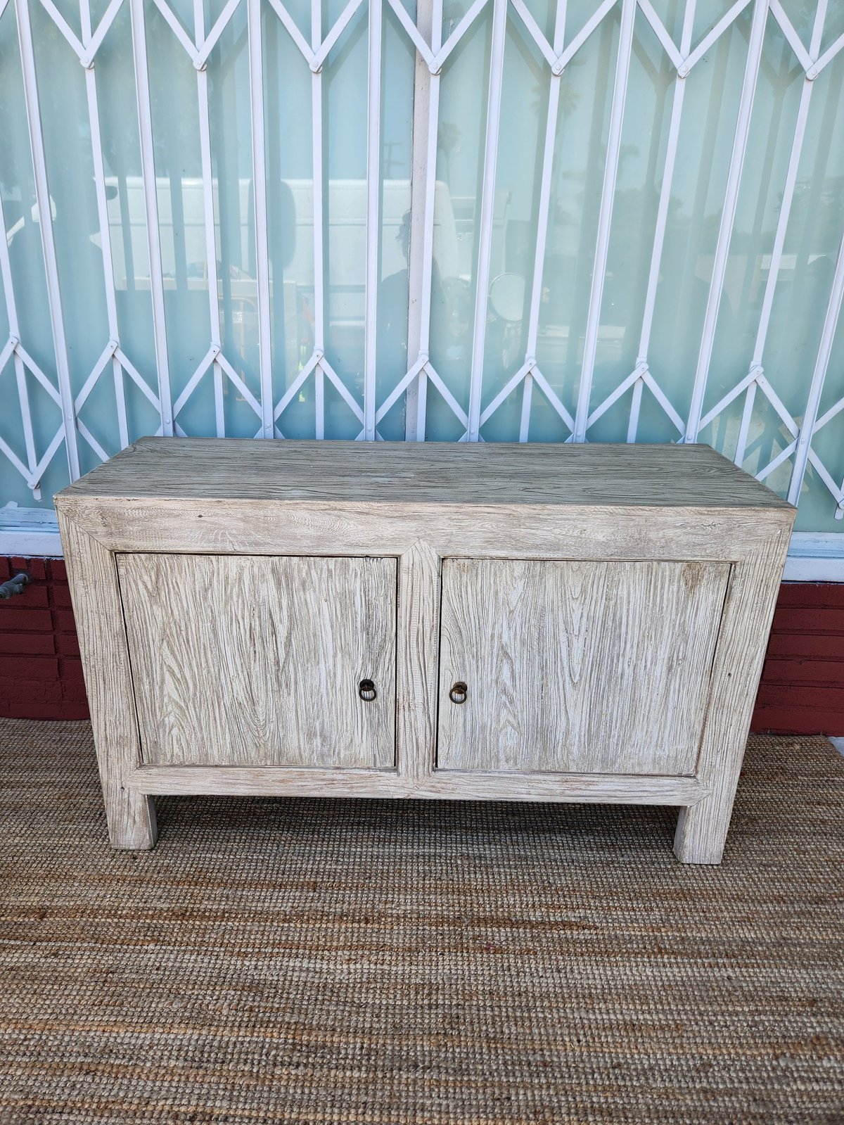 Whitewashed wooden cabinet with two doors and metal handles on a carpet, displayed in front of a patterned curtain