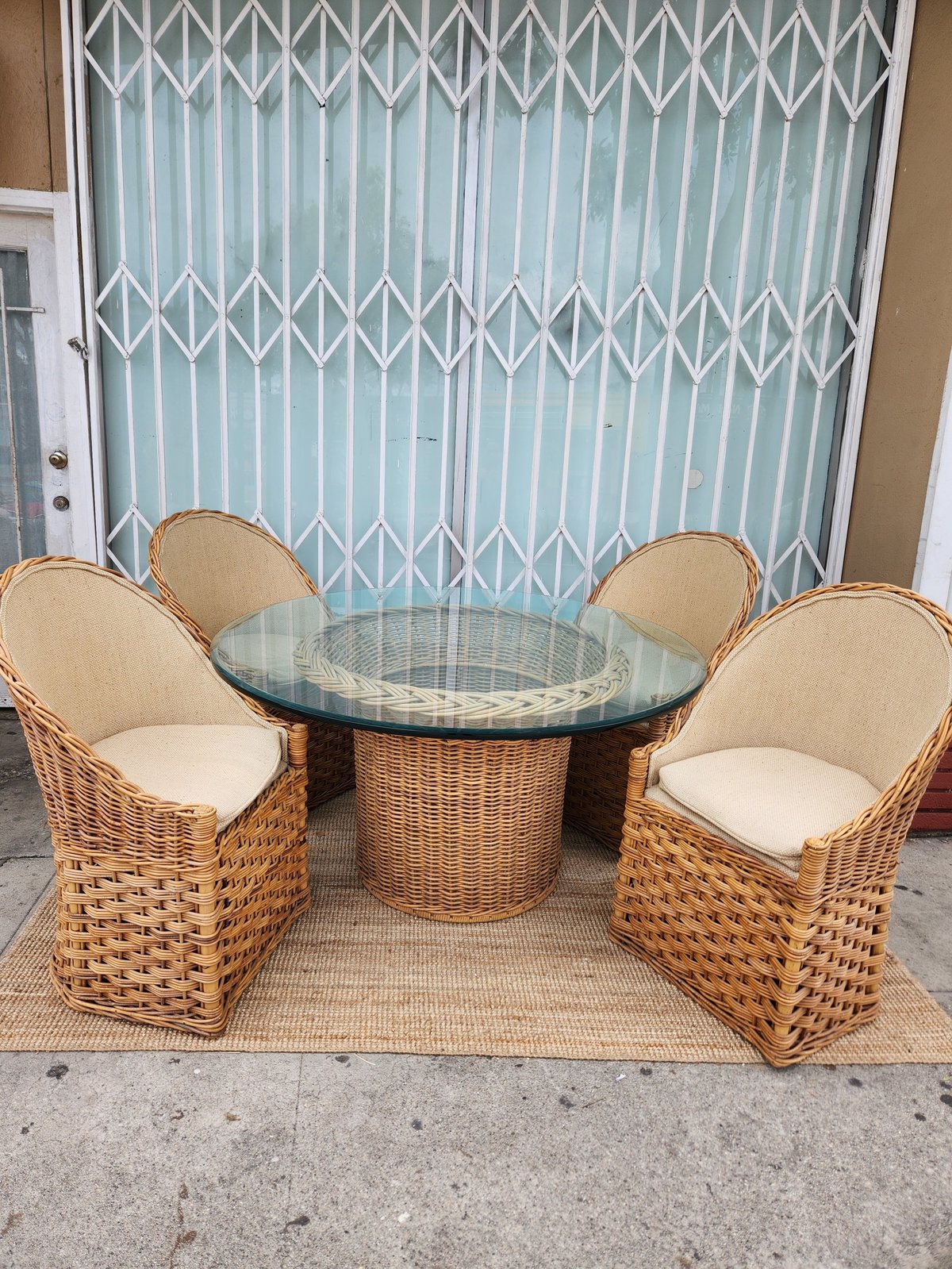 Wicker patio set with round glass-top table and three armchairs with cream cushions, positioned on a beige rug in front of a turquoise accordion door gate