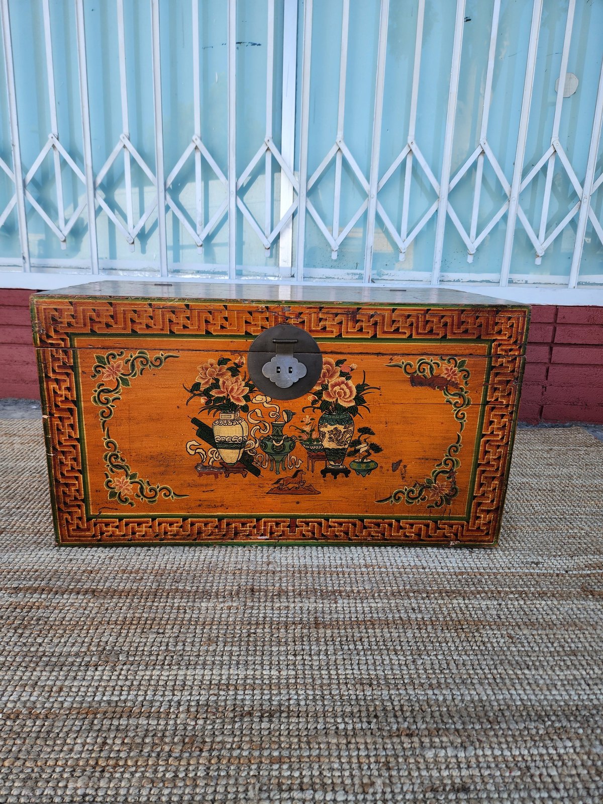 Ornate wooden chest with golden decorative designs and red geometric borders on a brick patio against a white fence