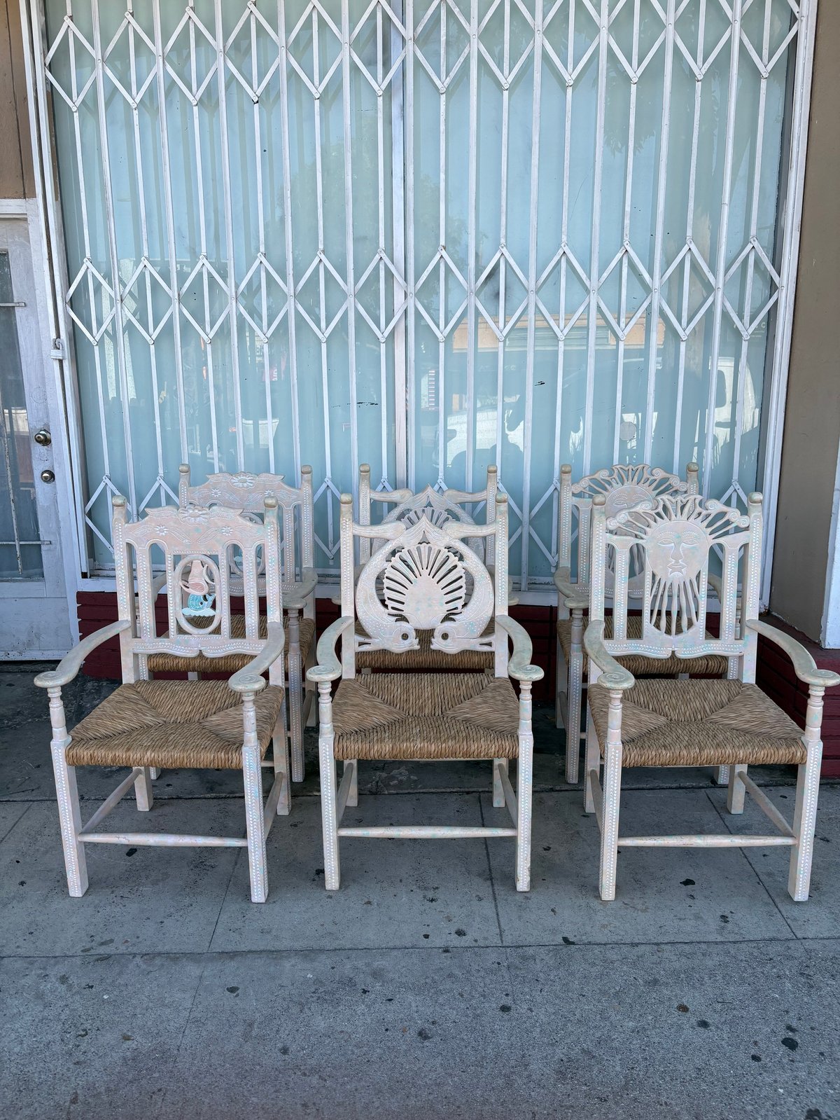 Three white wooden chairs with woven rush seats arranged in front of a light blue accordion security gate