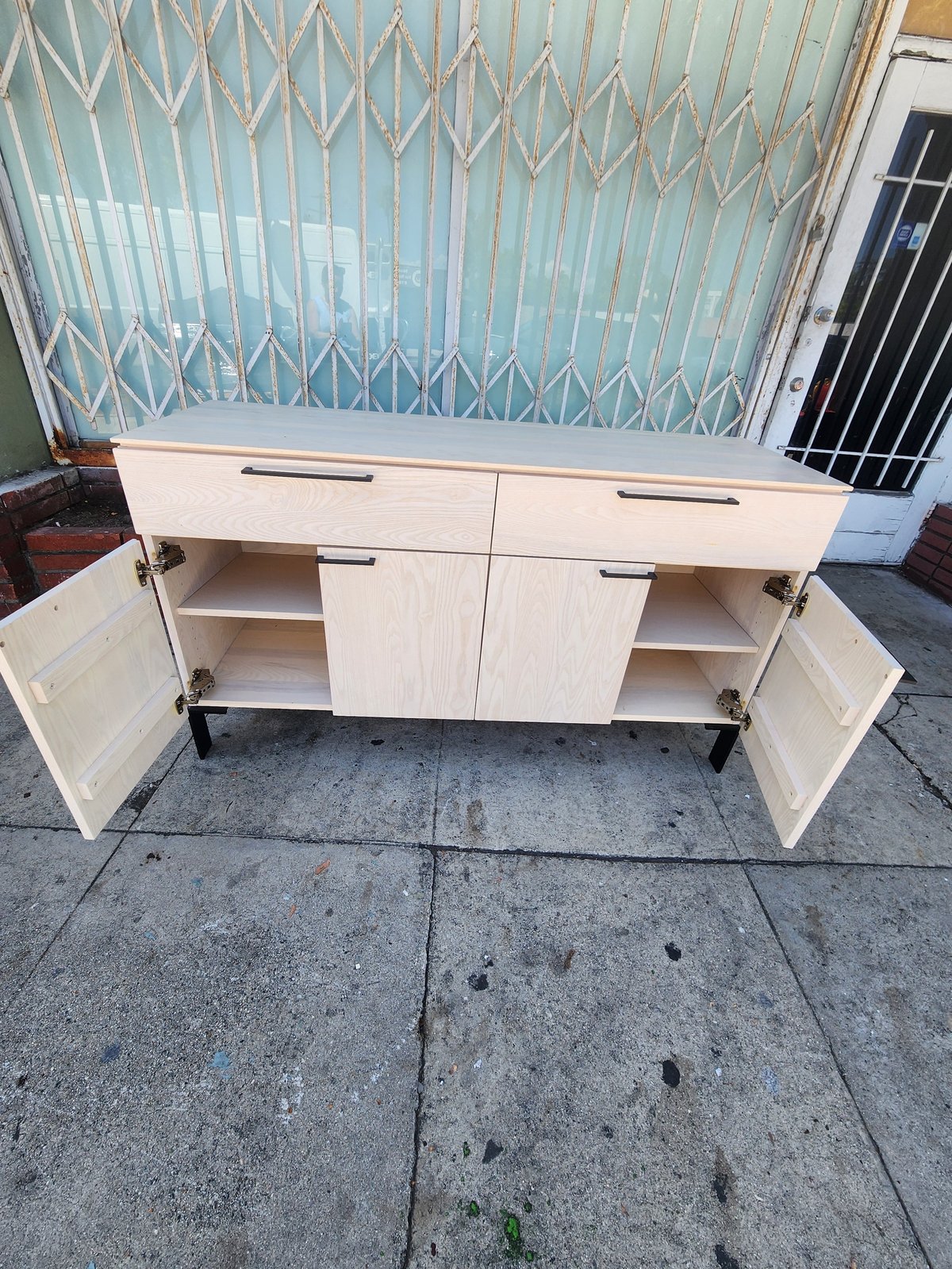 Light wooden cabinet with open doors and drawers displayed on outdoor pavement, showing interior shelving and storage space