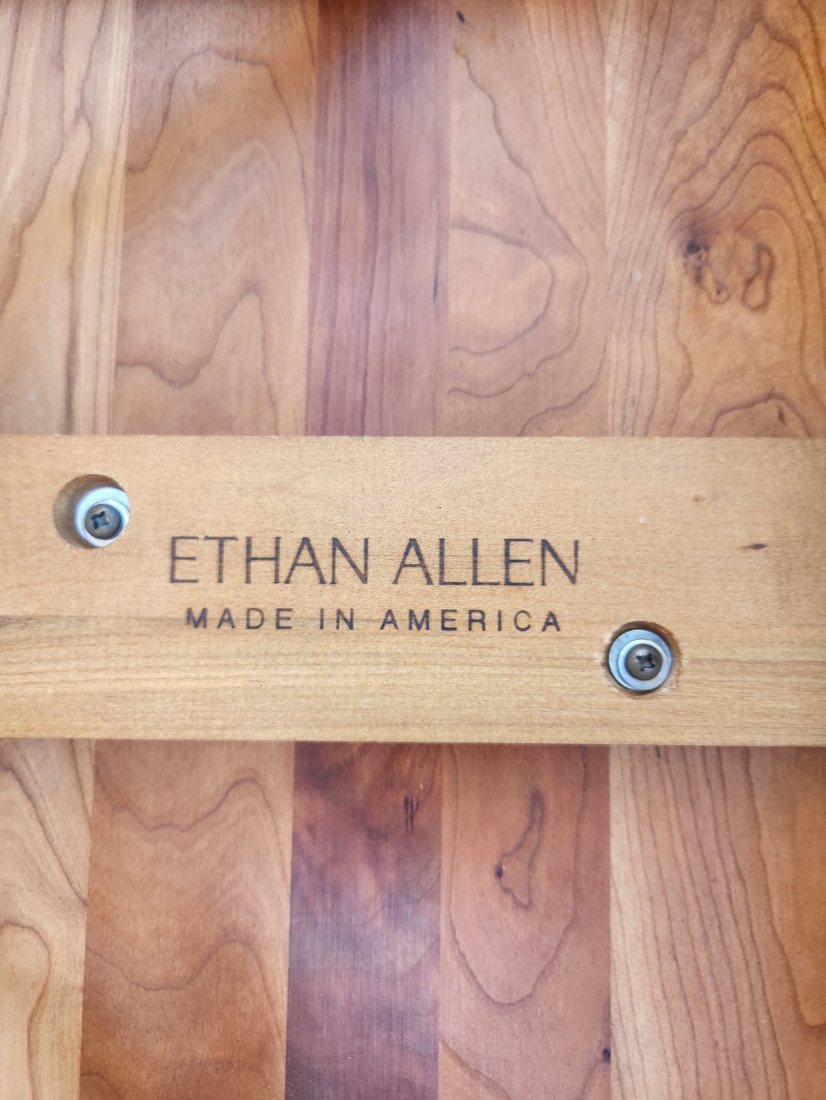 Wood nameplate reading ETHAN ALLEN MADE IN AMERICA with metal screws on walnut wood grain background