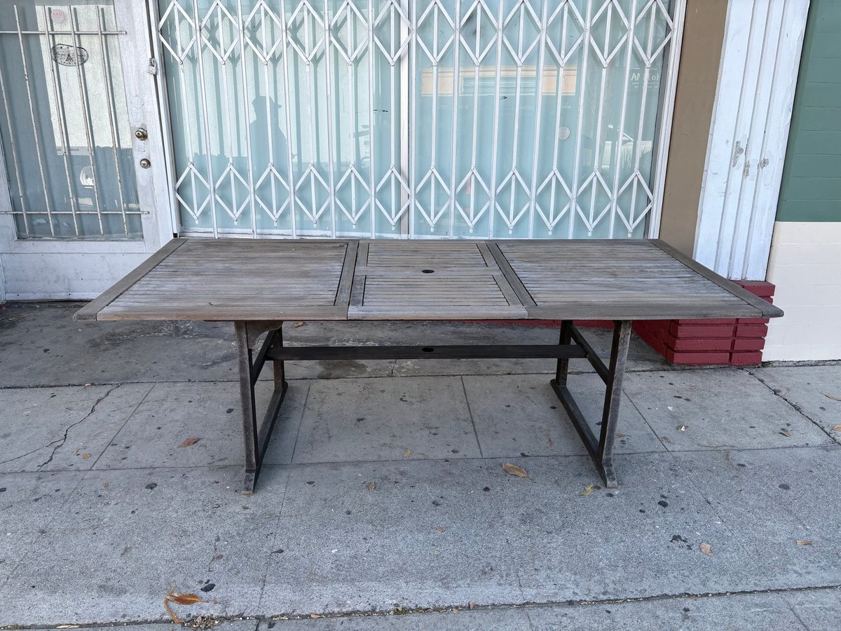 Weathered wooden patio table with metal frame on concrete ground in front of white metal gate and green door.