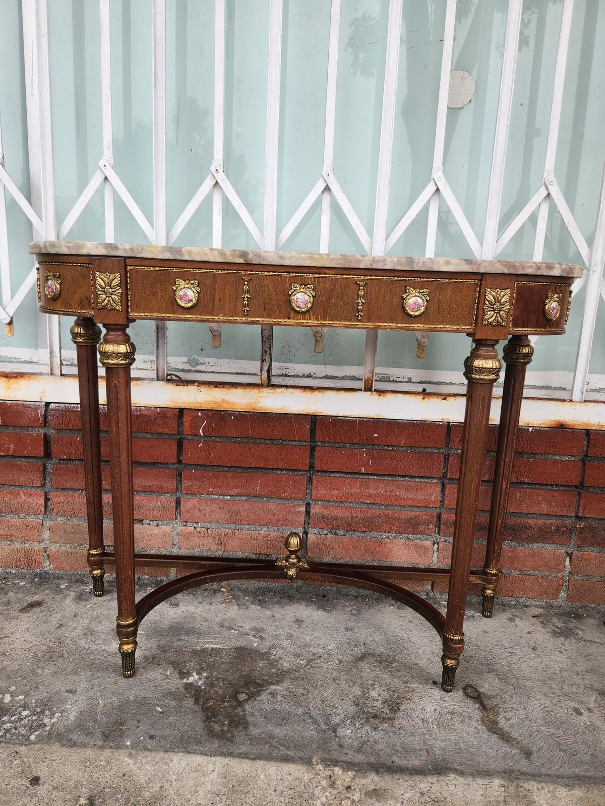 Antique wooden console table with decorative inlay and brass hardware against brick wall and corrugated metal backdrop