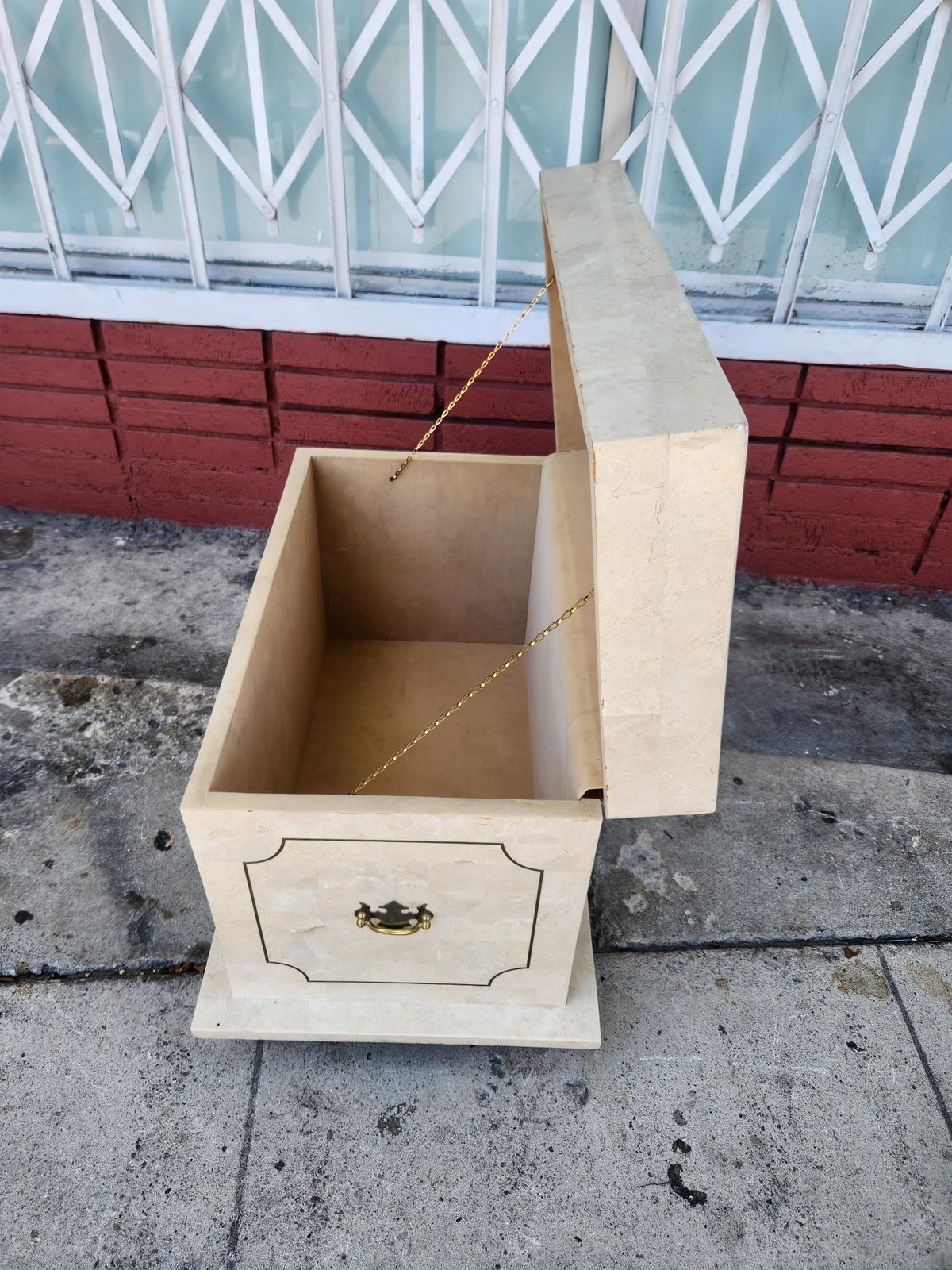 Unfinished wooden cabinet with open top drawer showing interior, brass hardware, on concrete ground with colorful fence behind