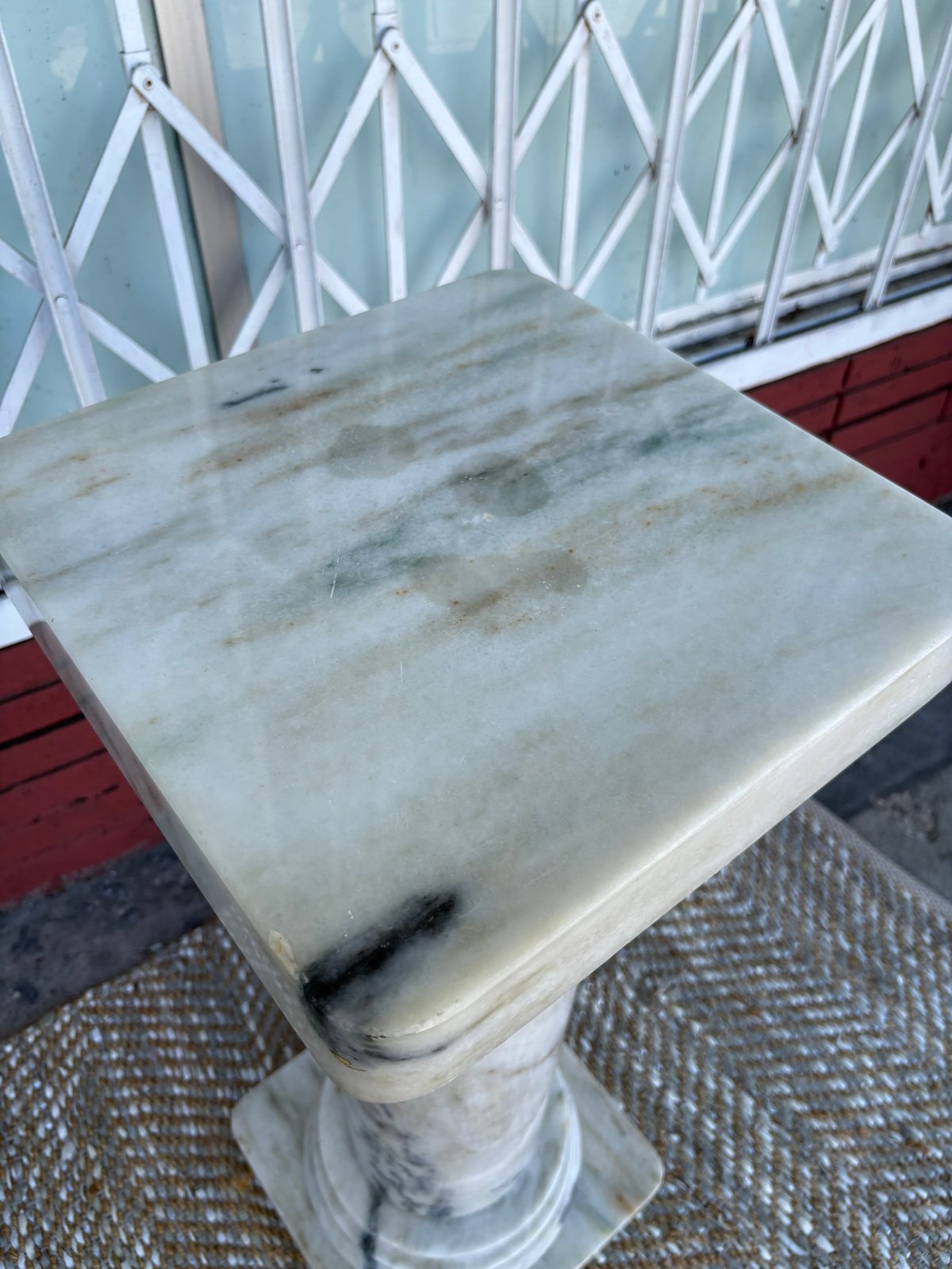 Weathered white marble pedestal table with stains on top, positioned on a deck with white lattice railings in background