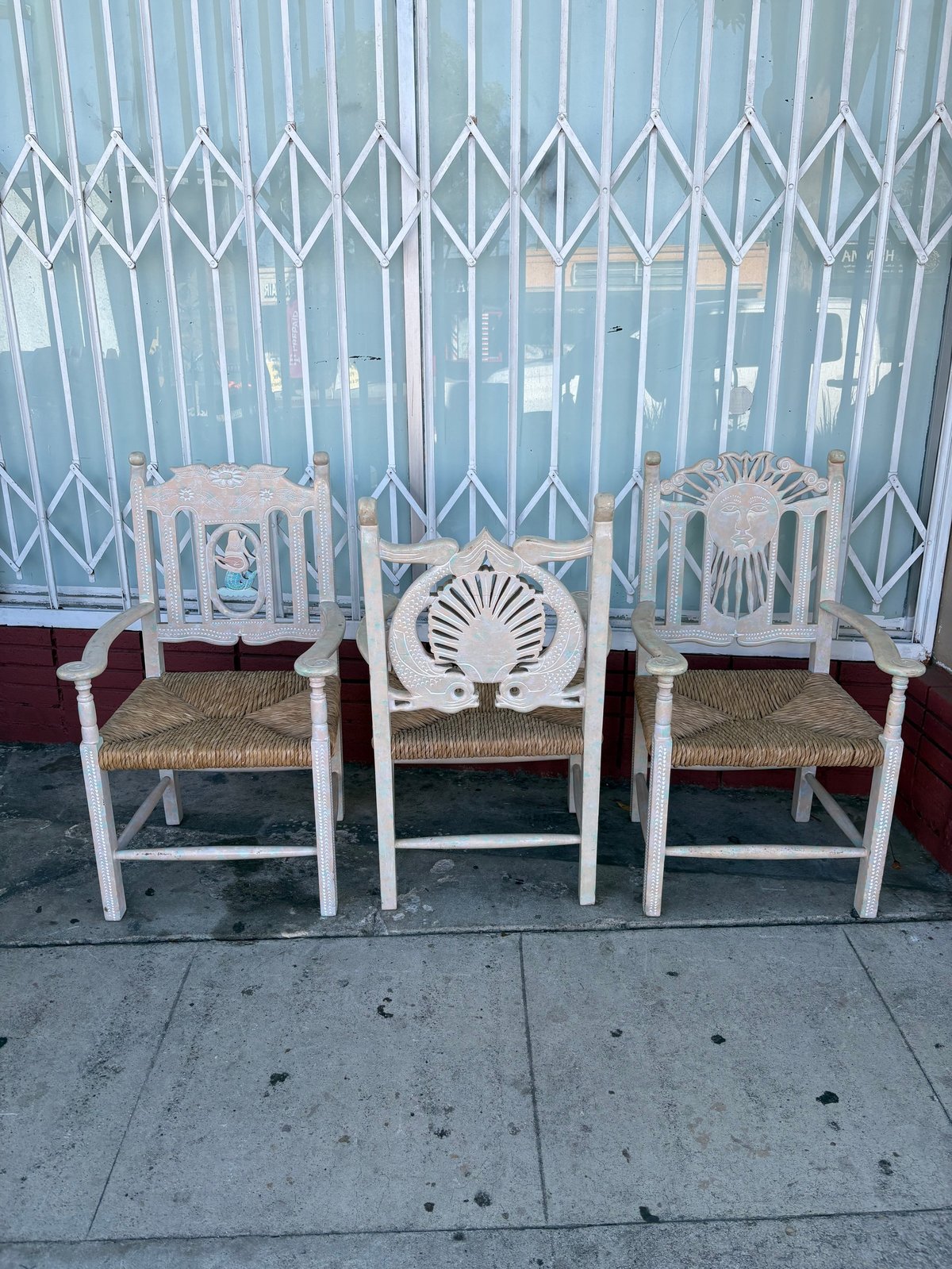 Three white wooden chairs with woven rush seats and decorative shell-shaped backs lined up against a metal accordion gate