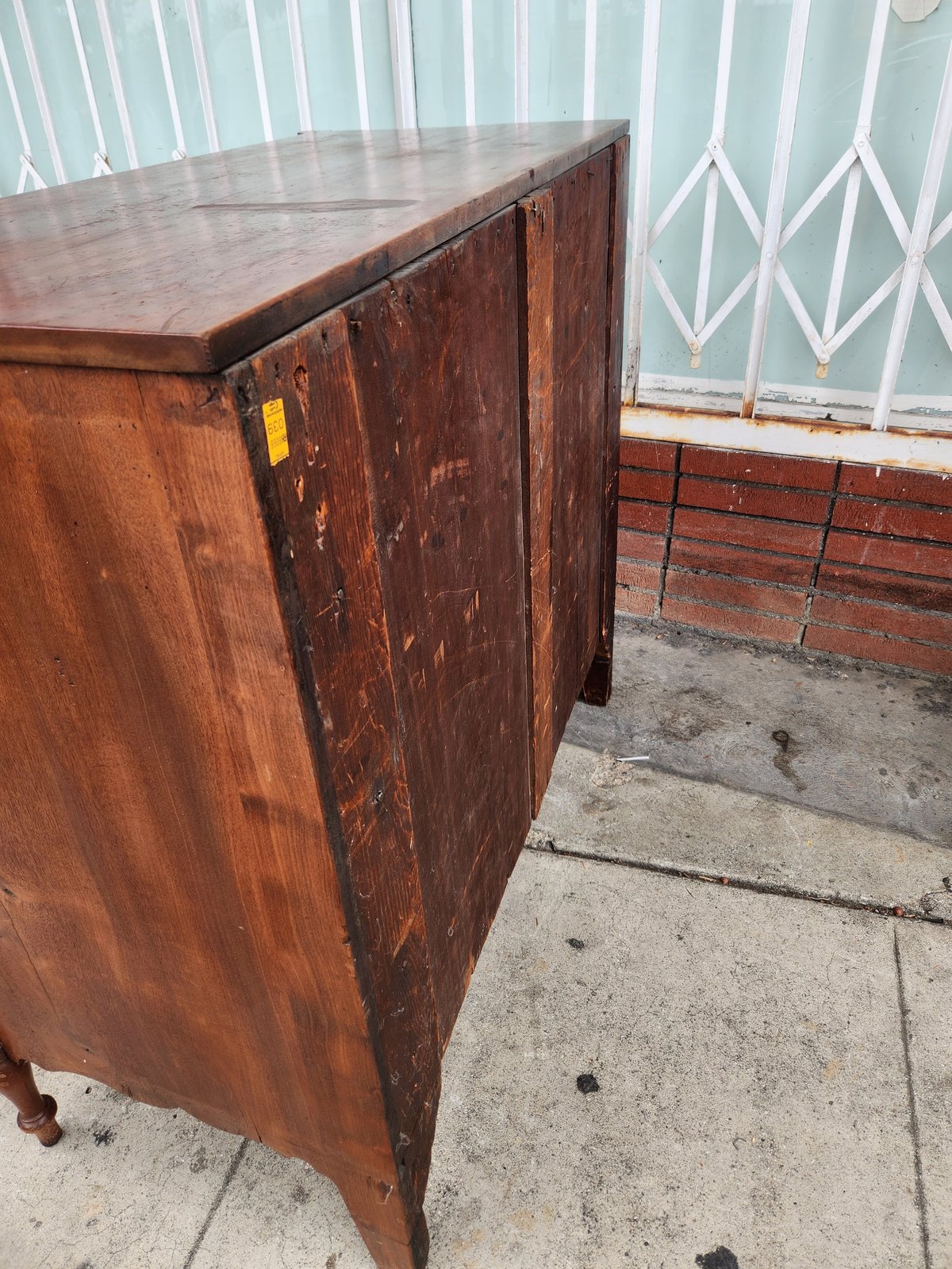 Vintage wooden cabinet with worn dark finish sitting on concrete beside brick wall and metal gate