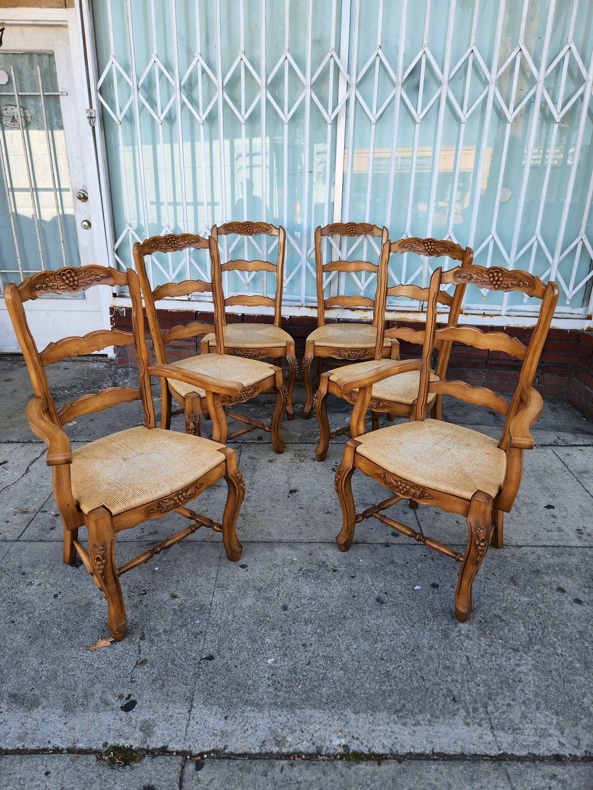 Six wooden dining chairs with woven rush seats arranged in a circle on a concrete surface against a corrugated metal garage door