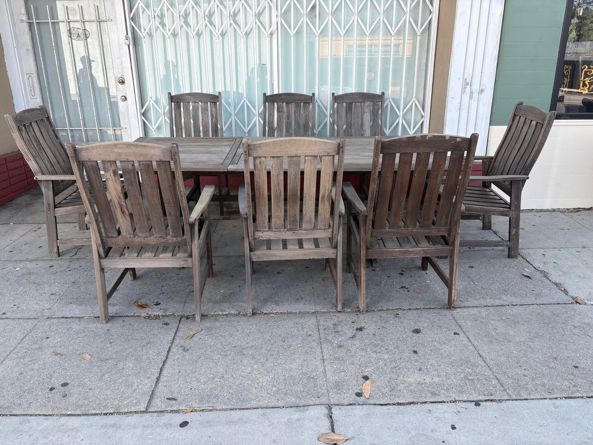 Outdoor patio furniture set with wooden dining table and six Adirondack chairs arranged on concrete patio near white gate and storefront.
