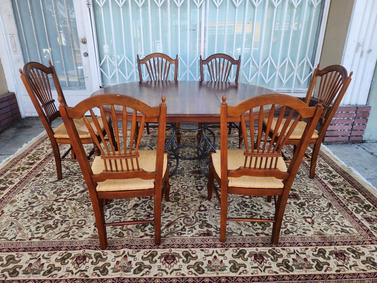 Wooden dining table with six shield-back chairs and cream-colored cushions on an ornate area rug in a bright room with patterned curtains