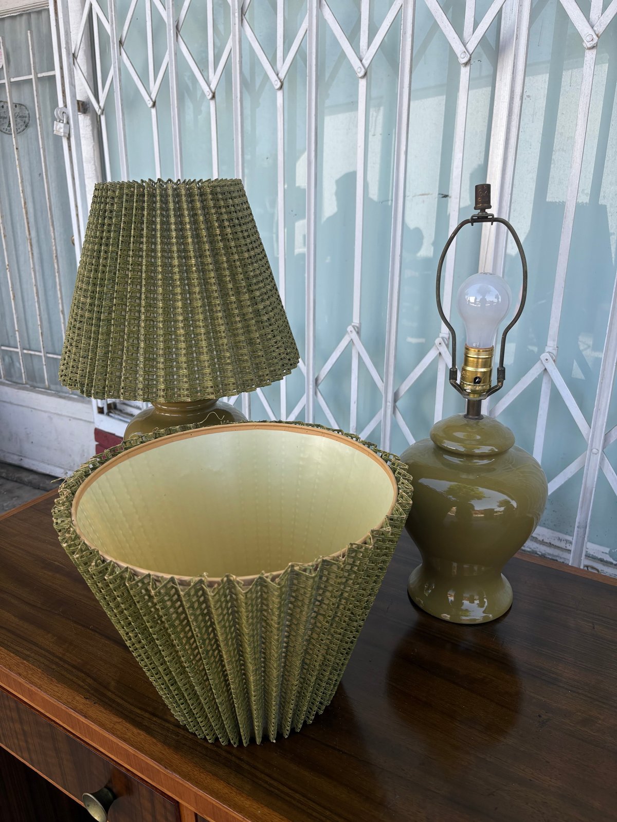Two olive green ceramic table lamps with pleated shades on a wooden dresser near a window with geometric white curtains