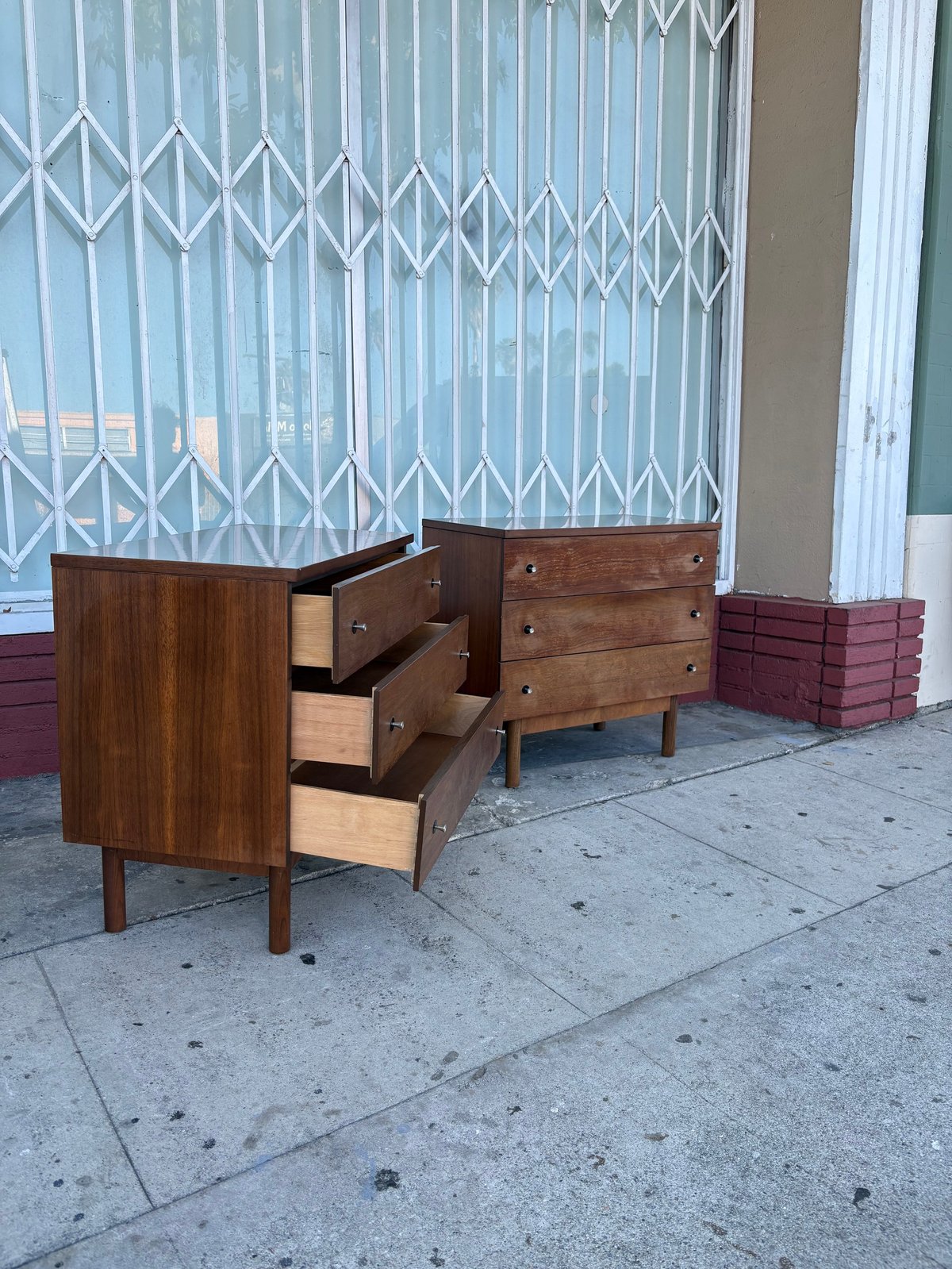 Two mid-century wooden dressers with open drawers displayed on a concrete sidewalk in front of a white metal security gate