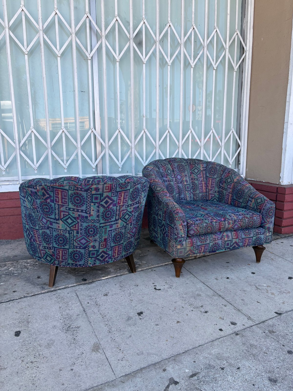 Two patterned armchairs with geometric designs on a sidewalk in front of an accordion gate and storefront