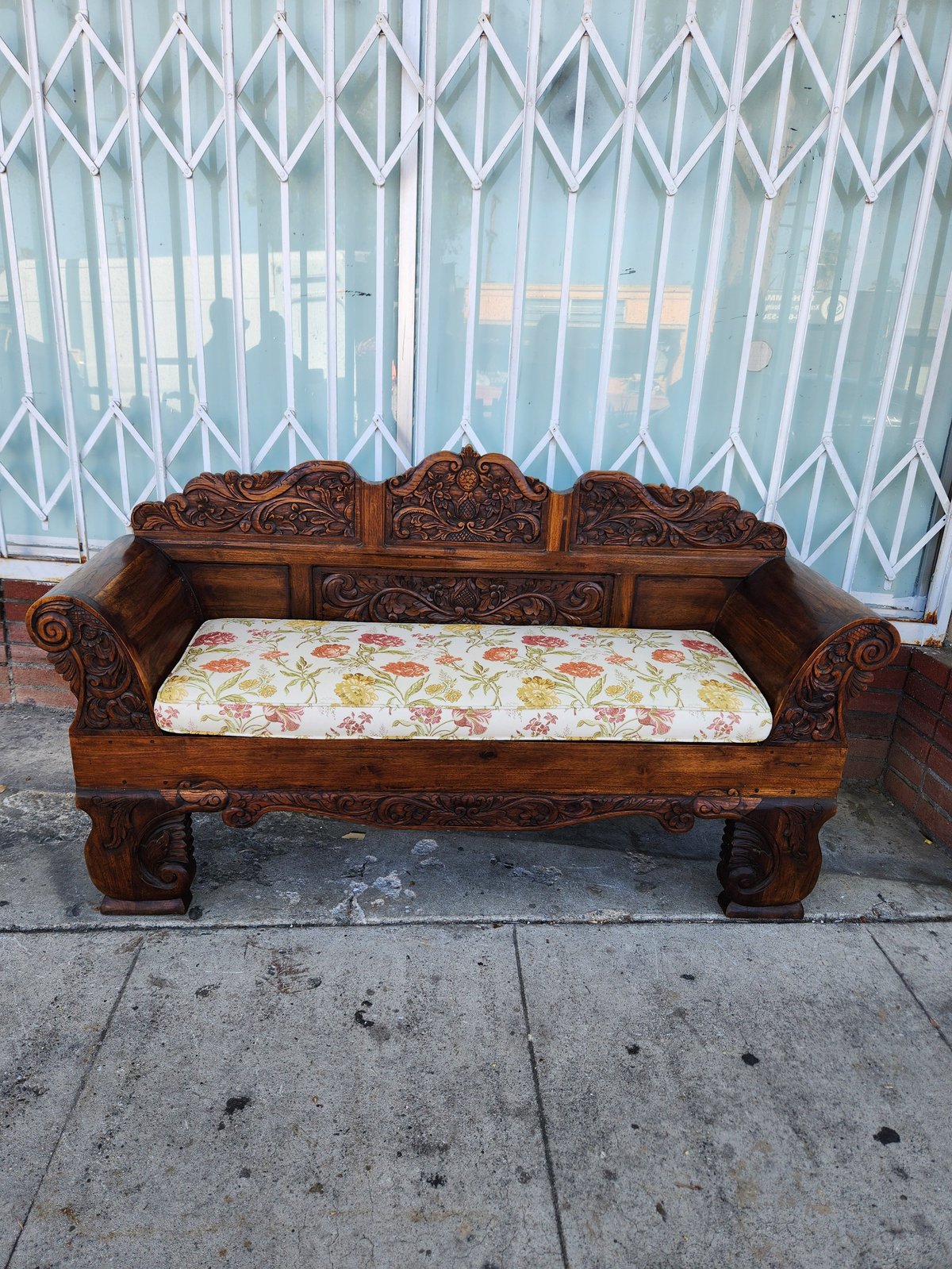 Ornately carved wooden bench with floral cushion displayed on concrete sidewalk in front of metal security gates