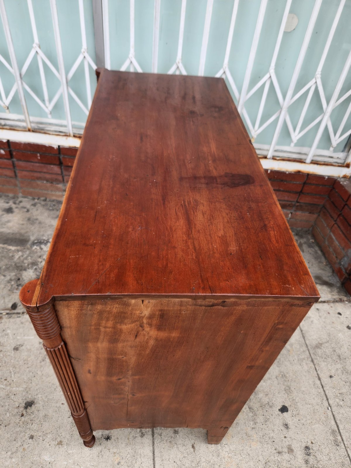 Vintage wooden dresser with rich reddish-brown finish and turned legs, photographed outdoors against a turquoise metal gate