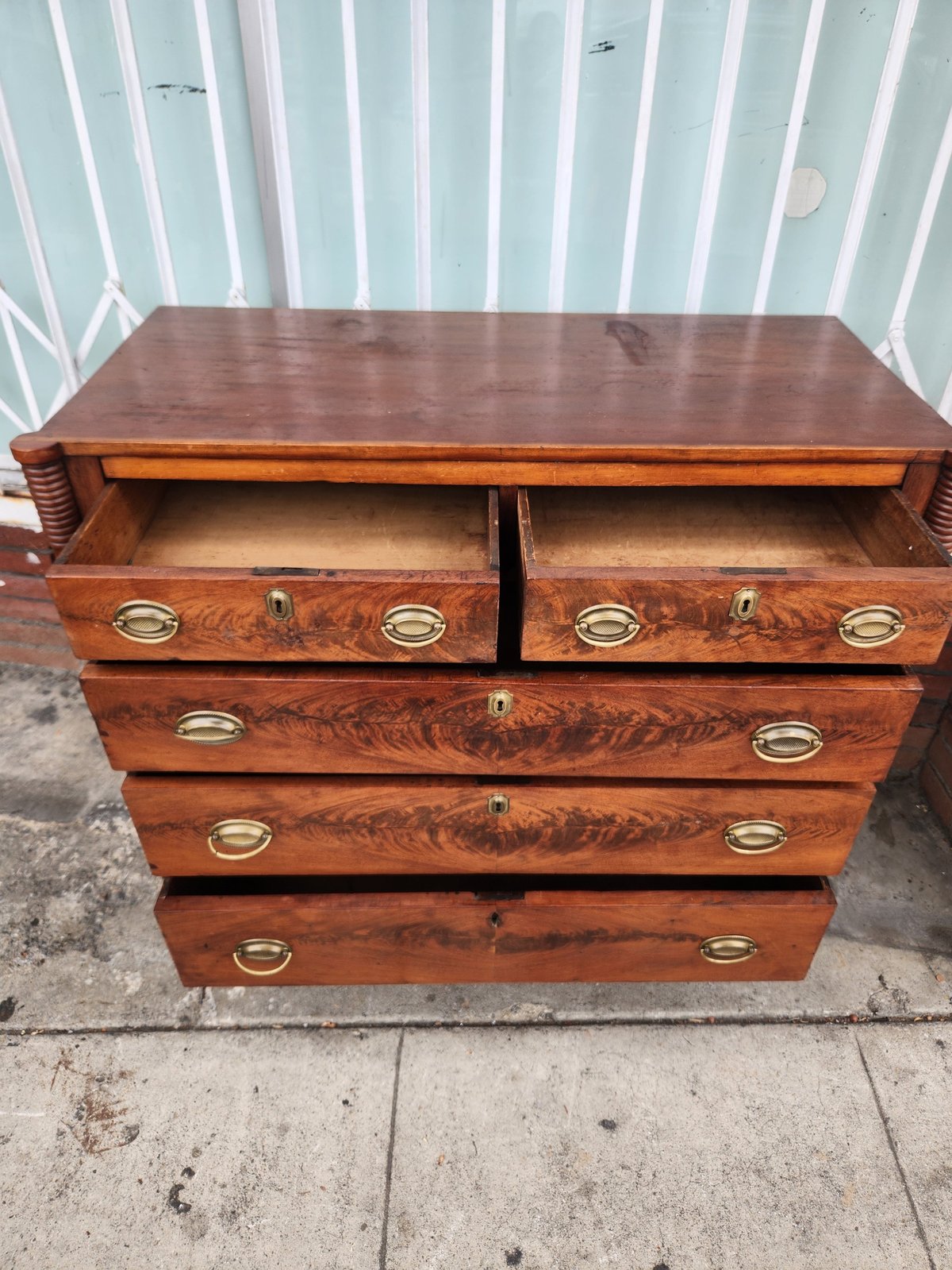 Wooden dresser with five drawers and brass hardware displayed outdoors against a corrugated metal wall