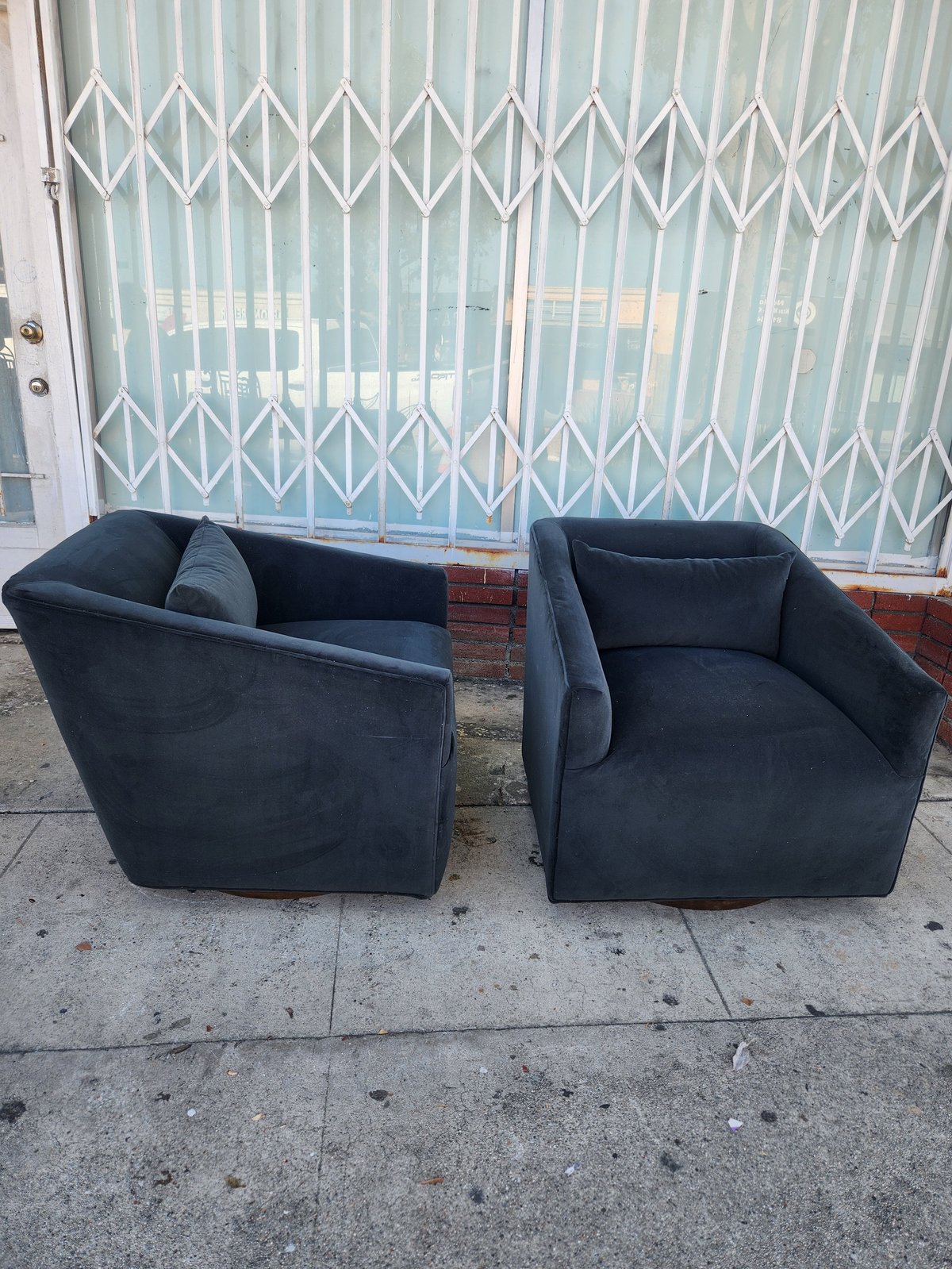 Two black curved armchairs on a concrete sidewalk in front of an accordion-style metal gate