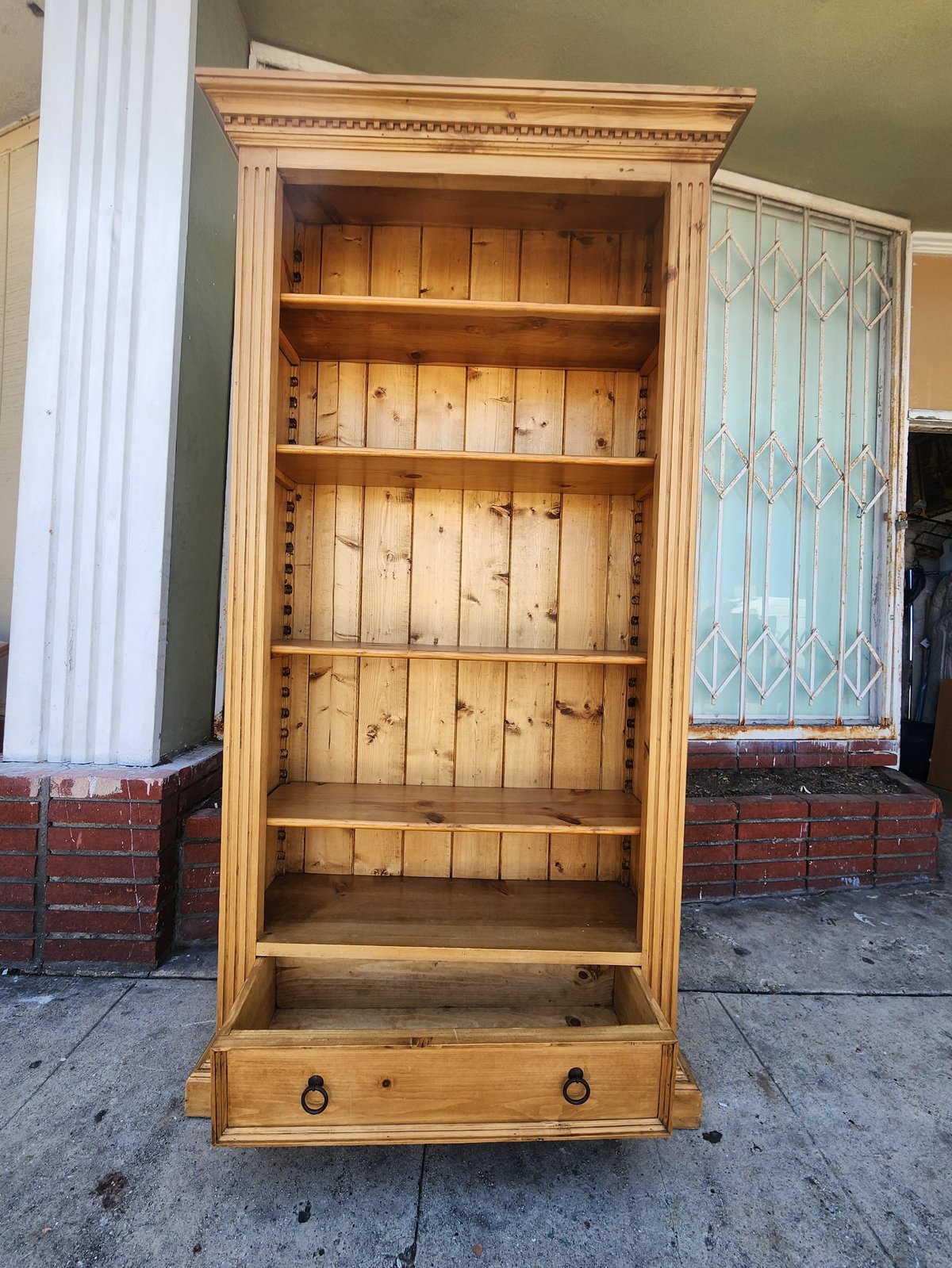 Wooden bookcase with decorative molding, five shelves, and a drawer at the base, displayed outdoors on concrete