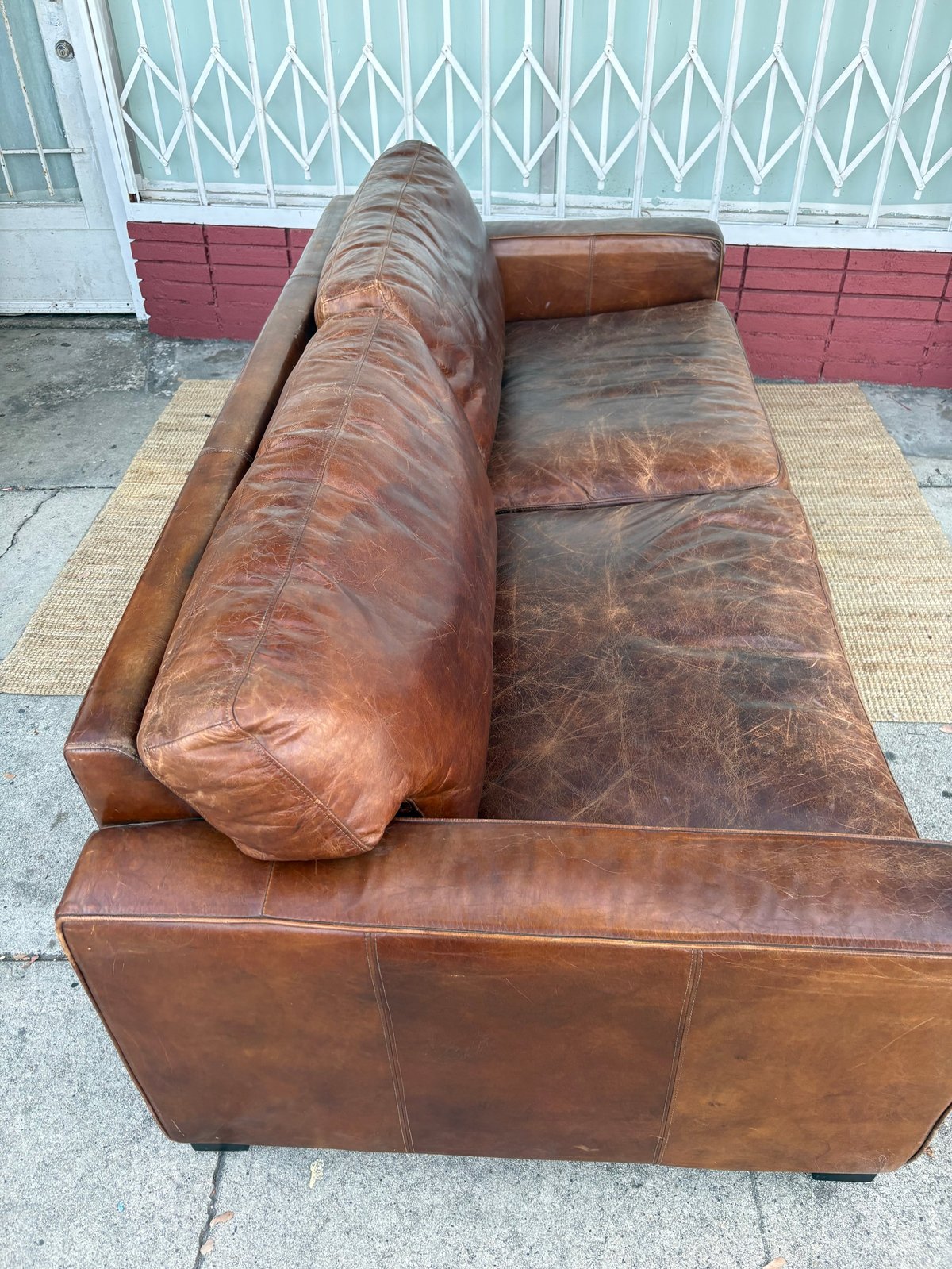 Worn brown leather sofa with bolster pillow on concrete driveway in front of white metal gate and red brick wall