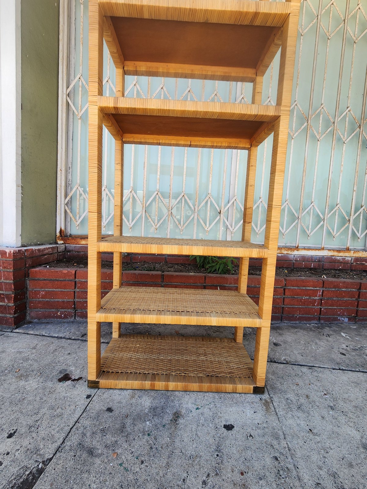 Wooden shelving unit with four woven rattan shelves displayed outdoors against metal gates and brick wall